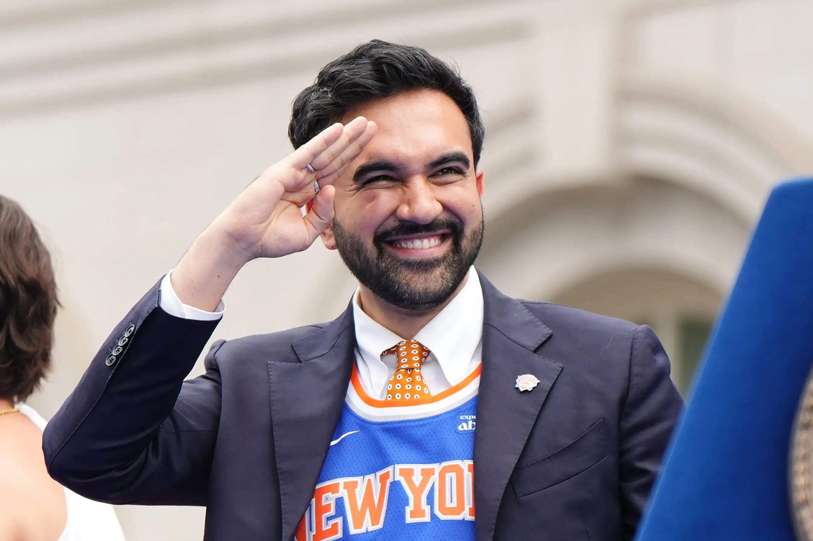 Zohran Mamdani smiles and salutes while wearing a NEW YORK basketball jersey beside a lectern with a city seal.