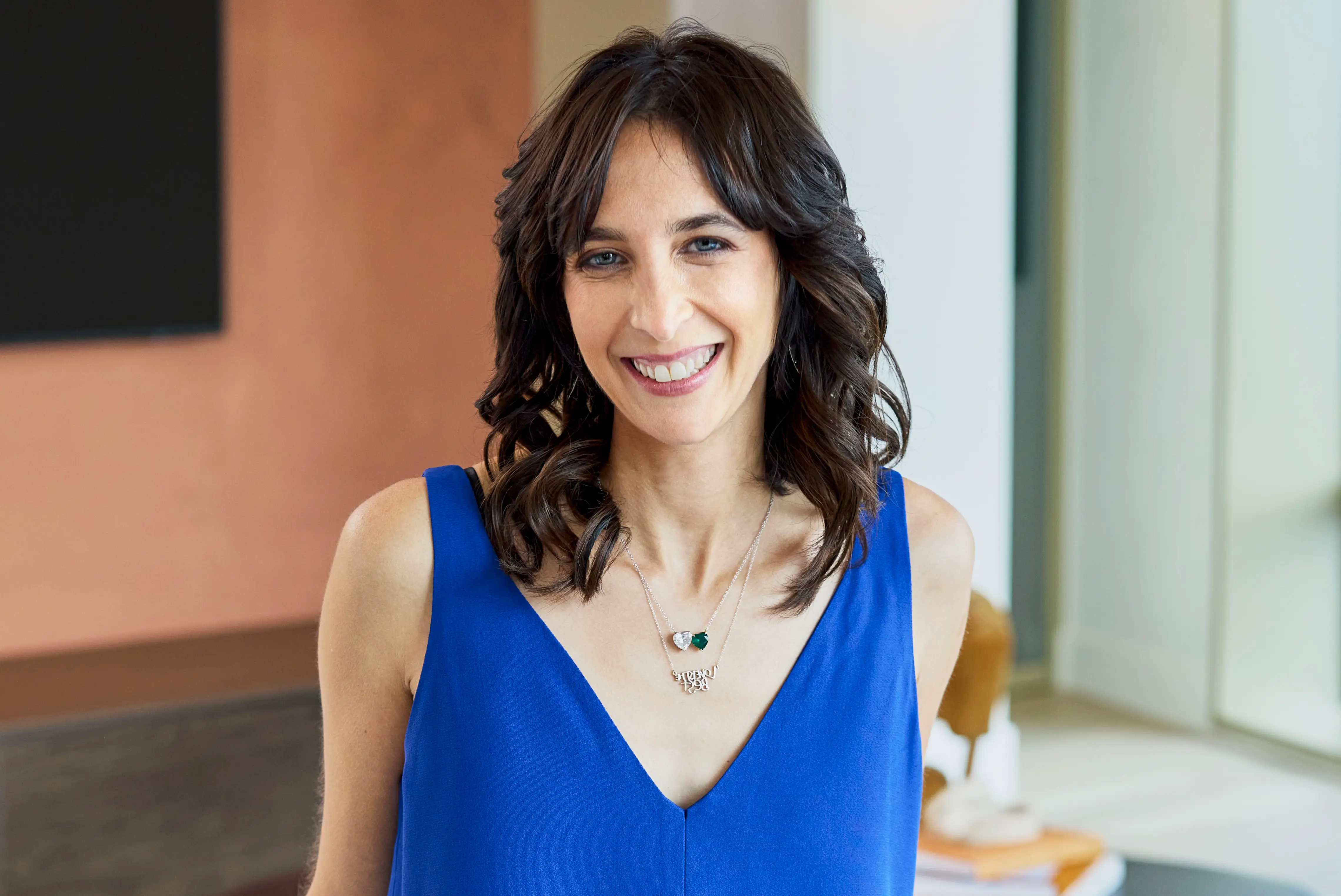 Smiling person wearing a bright blue sleeveless top stands indoors near large windows.