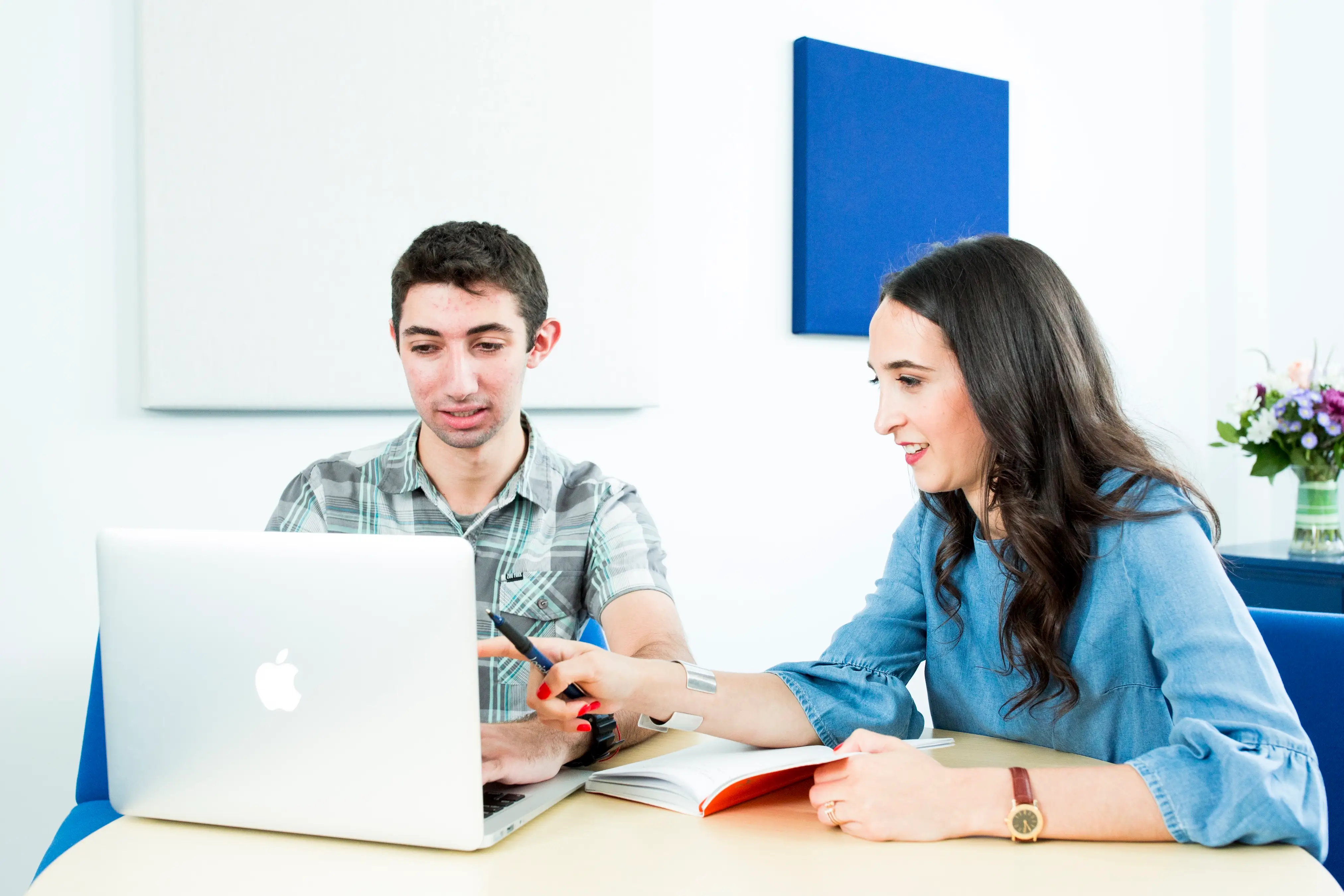 Two people work at a table with an Apple laptop, notebook, and pen in a bright office setting.