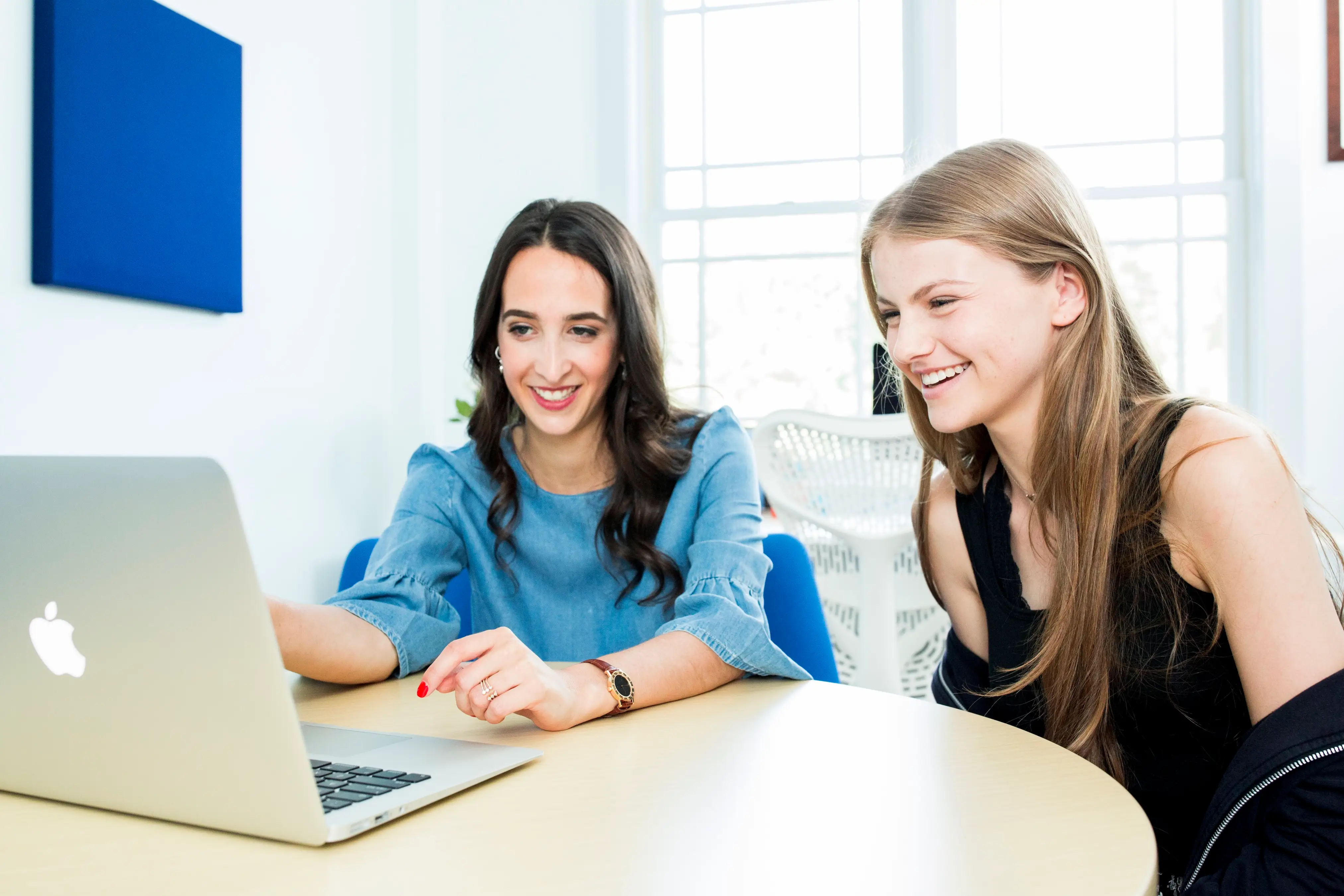 Two people smile while looking at an Apple laptop on a round table in a bright office.