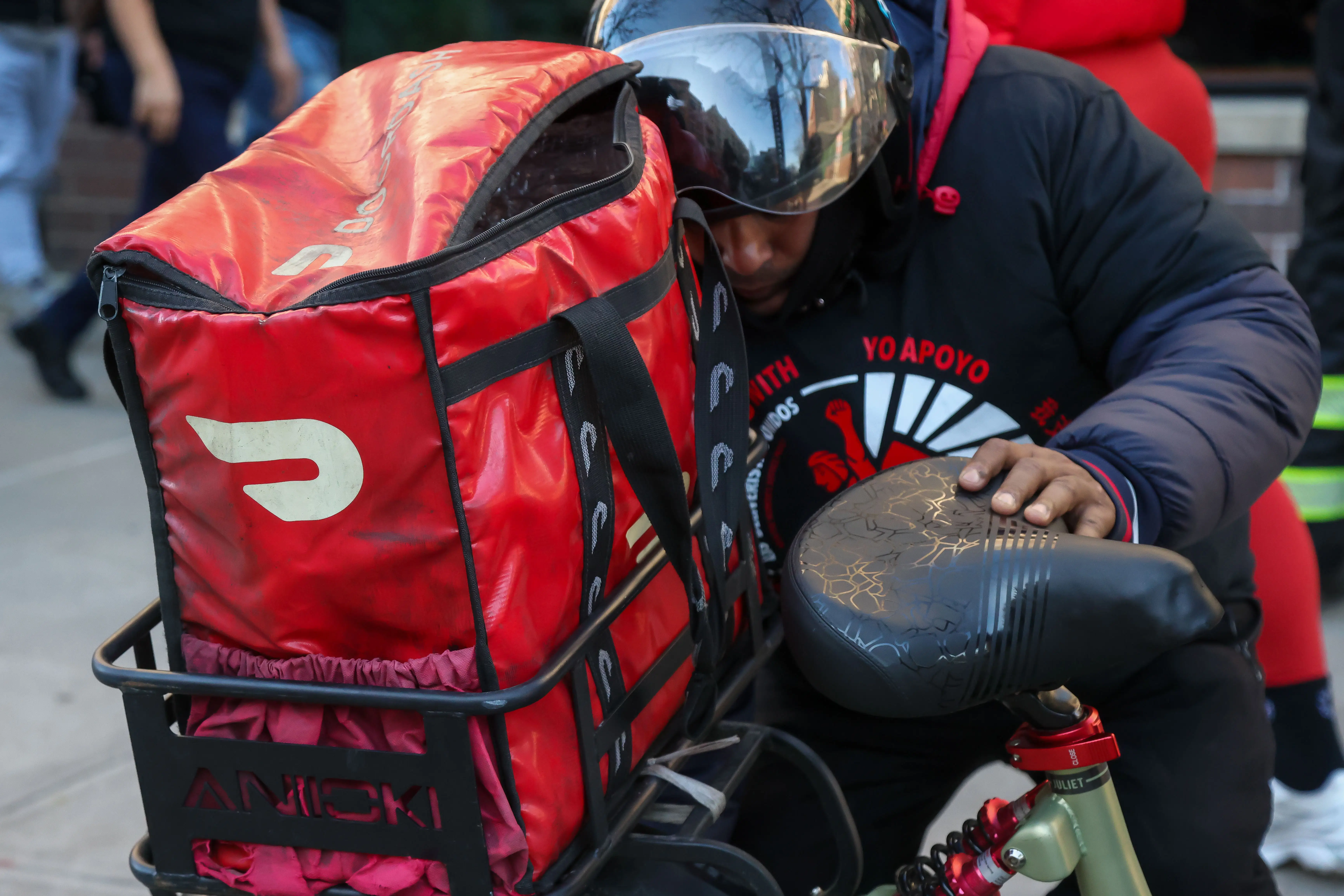 A Doordash delivery worker stands next to a bike while wearing a helmet. In a metal basket sits a red insulated DoorDash bag.