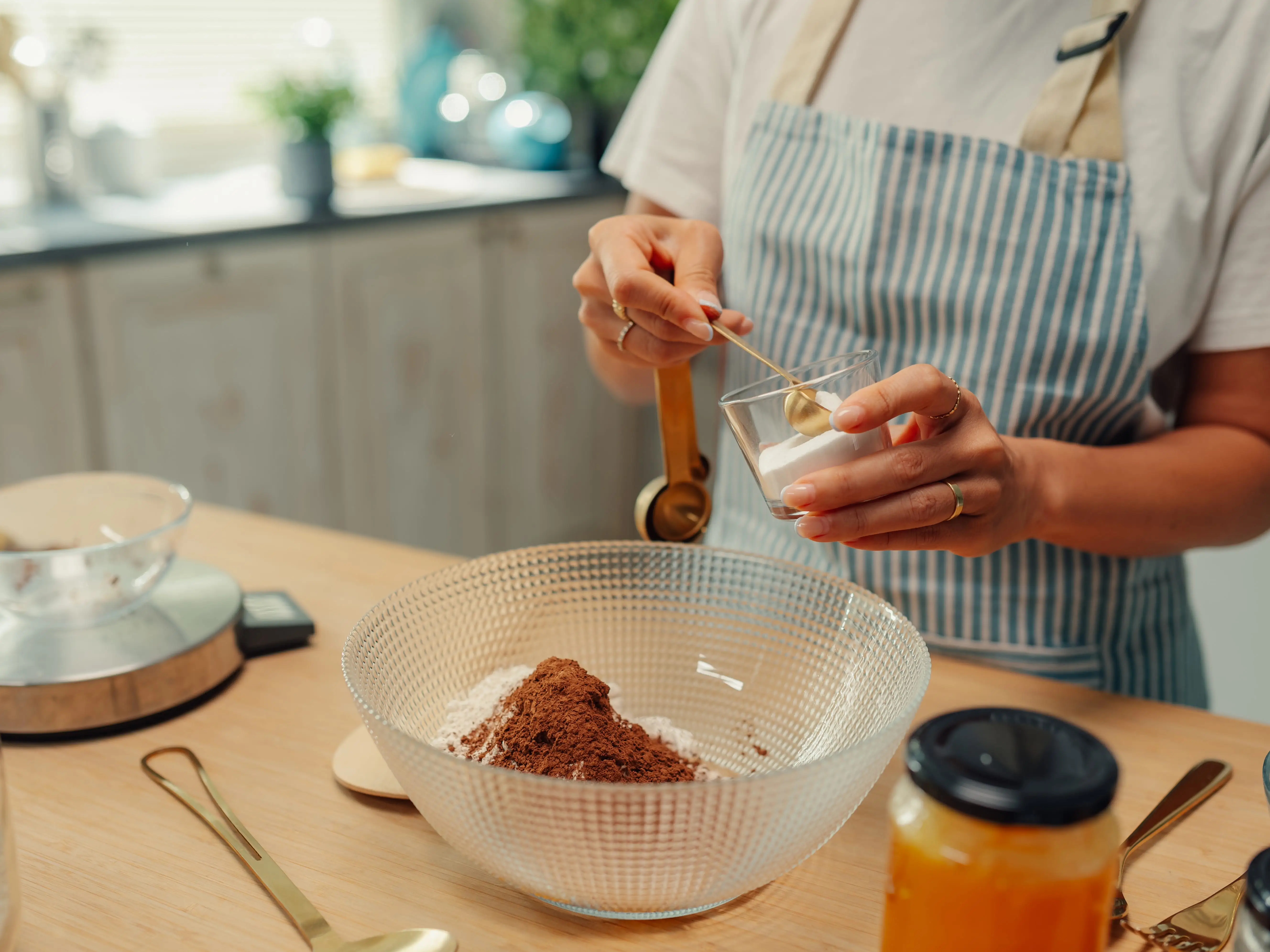 A person adding baking soda to a glass mixing bowl filled with other dry ingredients.