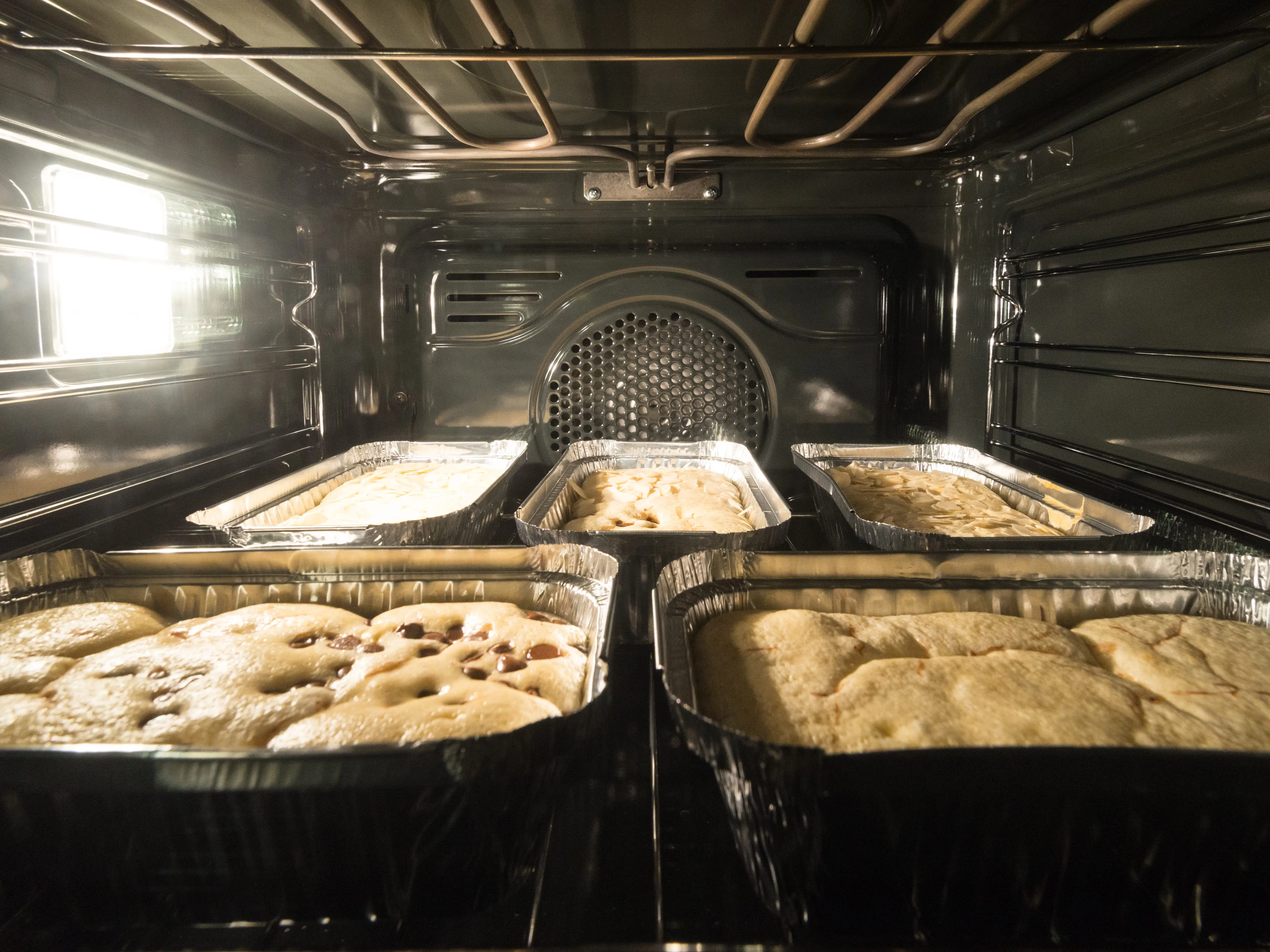 Several aluminum pans of baked goods sit on racks inside a lit oven.