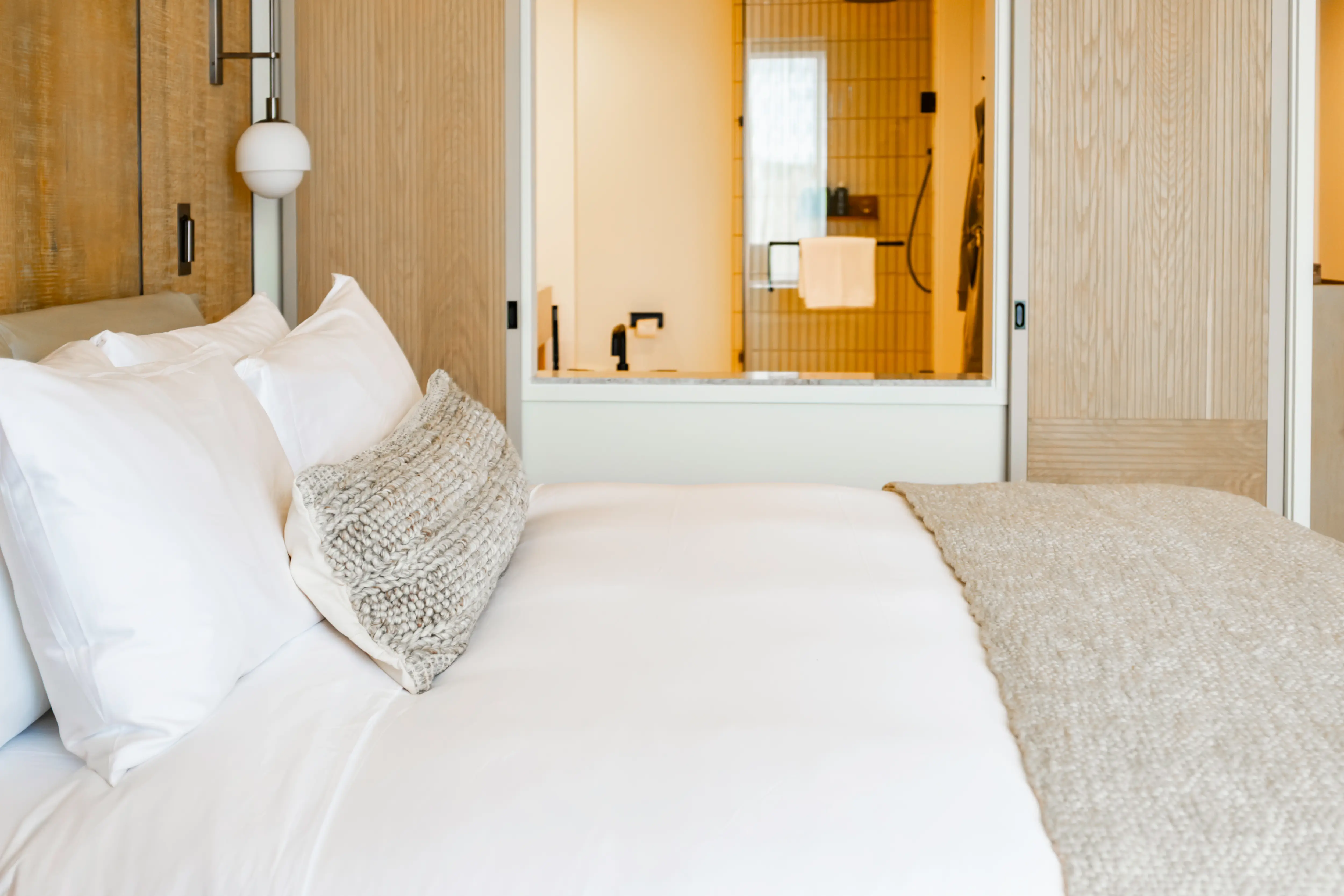 Modern bedroom with white bedding, textured throw pillow, wood paneling, and a bathroom visible through glass.