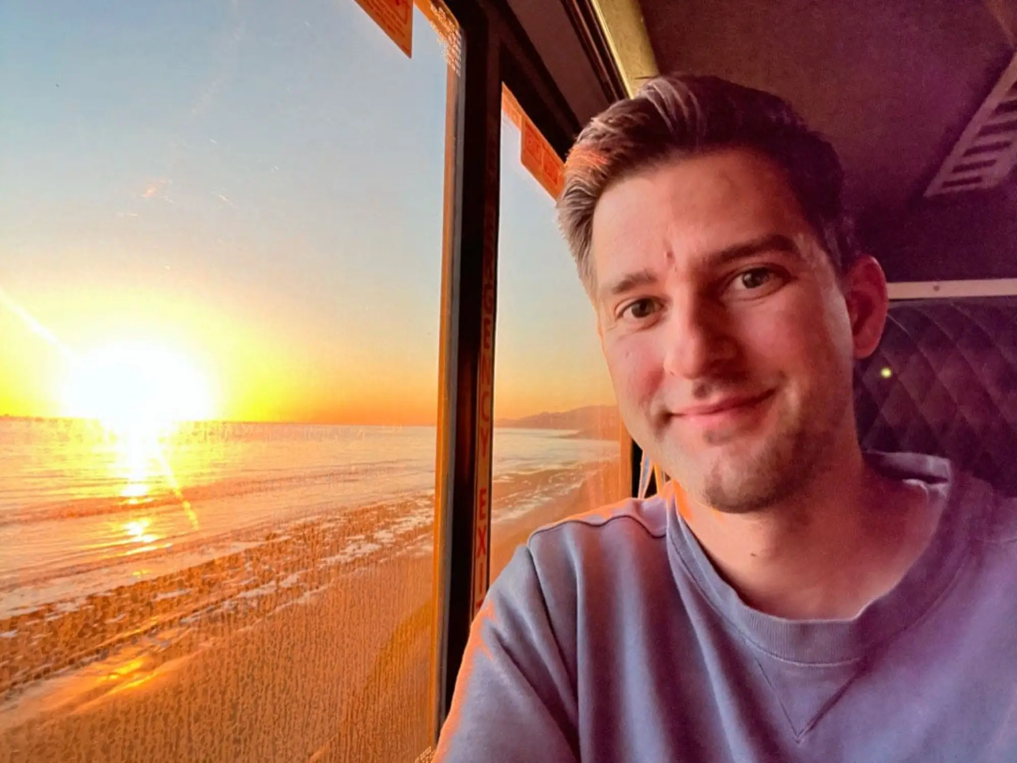 William takes a selfie near a train window with a view of the beach at sunset.