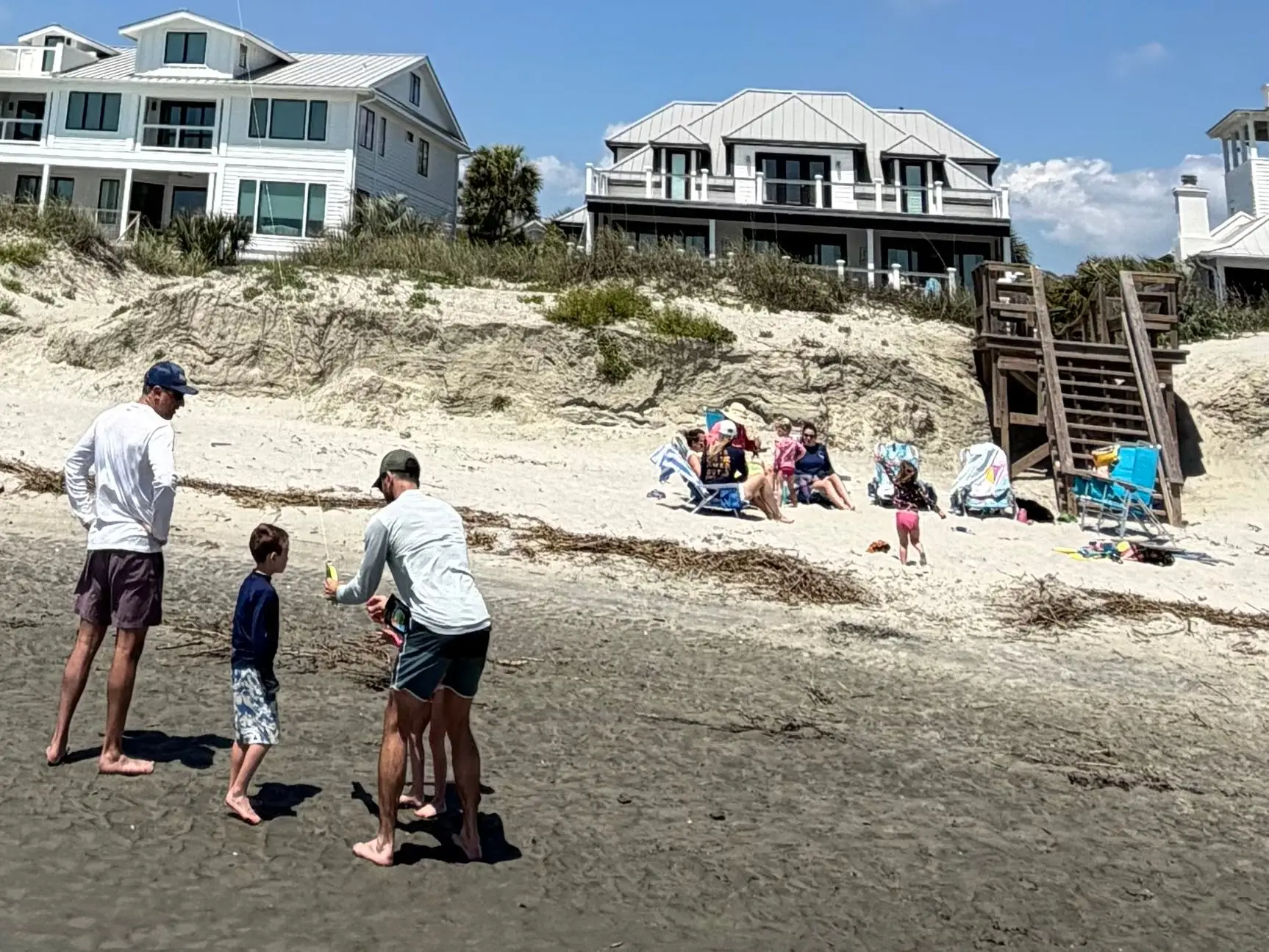 Families relax and play on the sand near a large beach house.