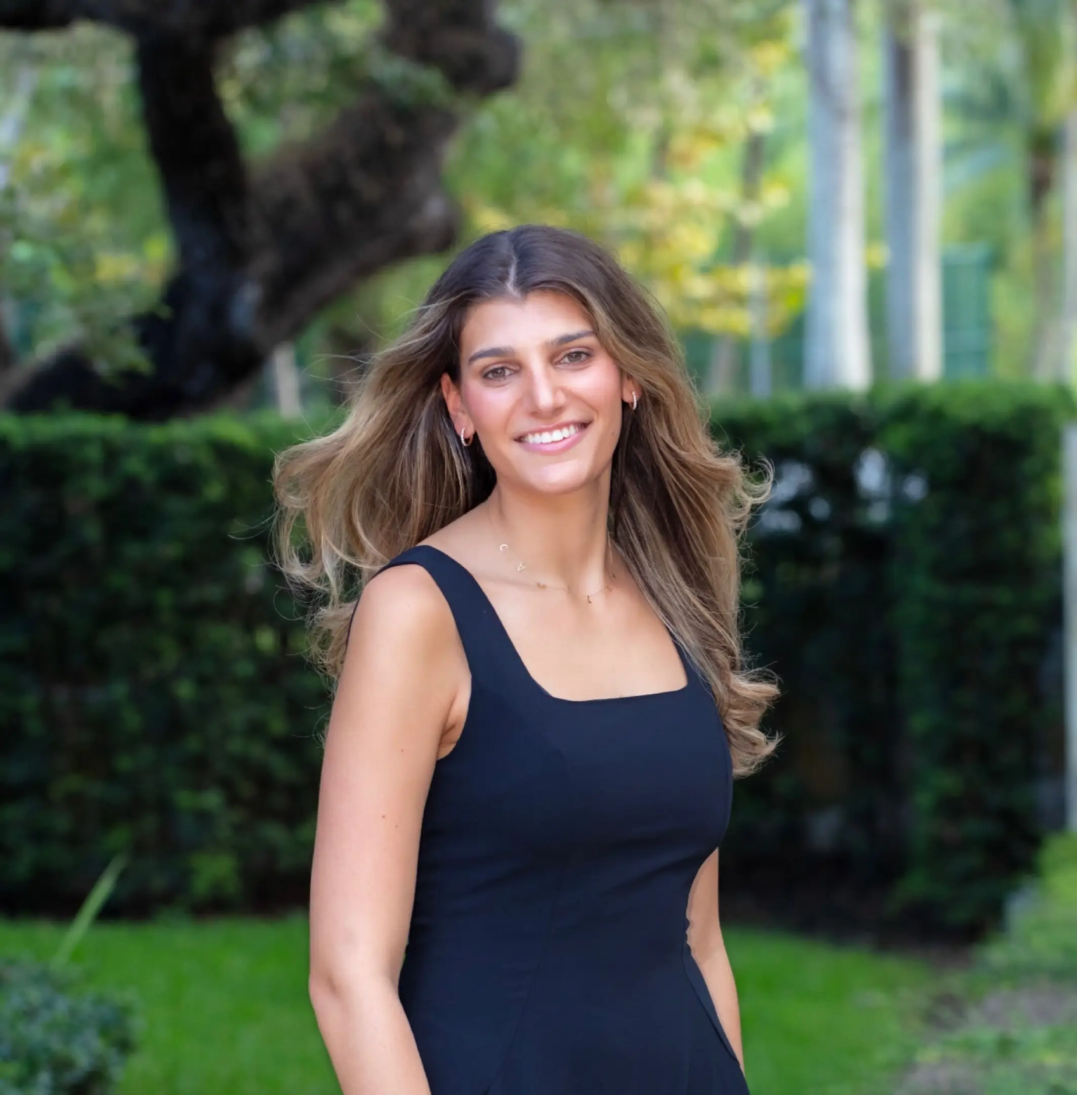 Smiling person in a black sleeveless dress stands outdoors in a green garden setting.