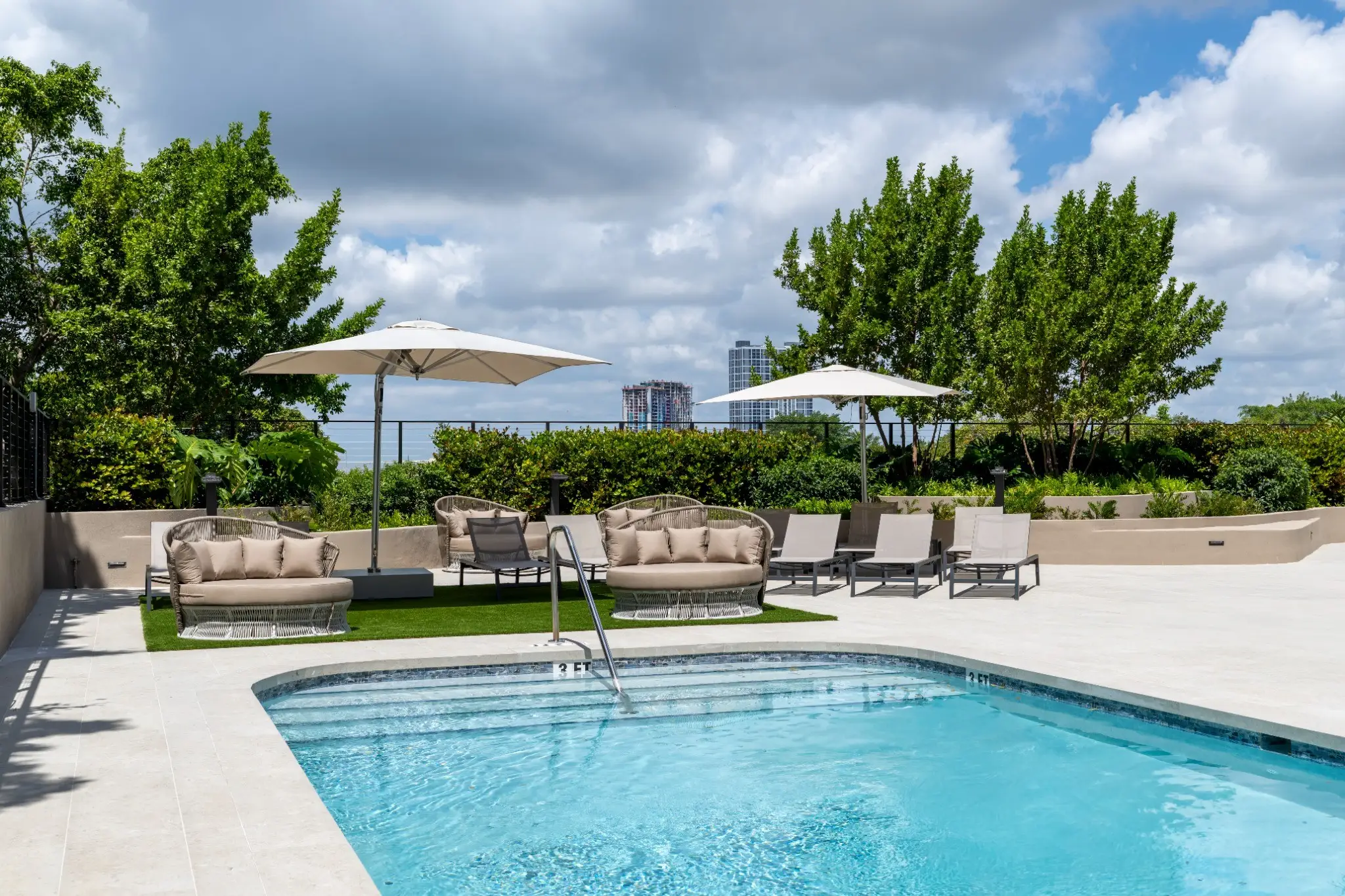 Outdoor pool deck with lounge seating, white umbrellas, greenery, and distant high-rise buildings under a cloudy sky.