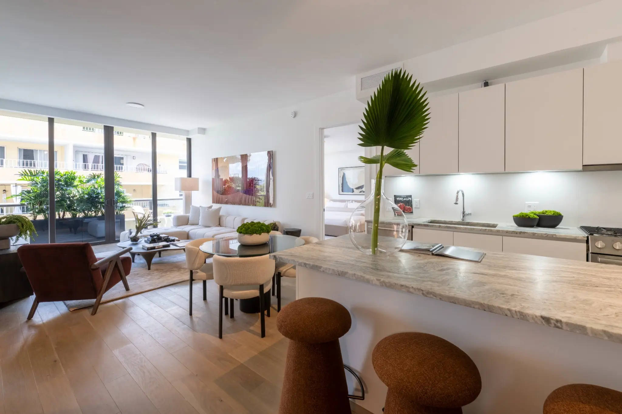Open-plan apartment interior with kitchen island, bar stools, dining area, living room, and balcony doors.
