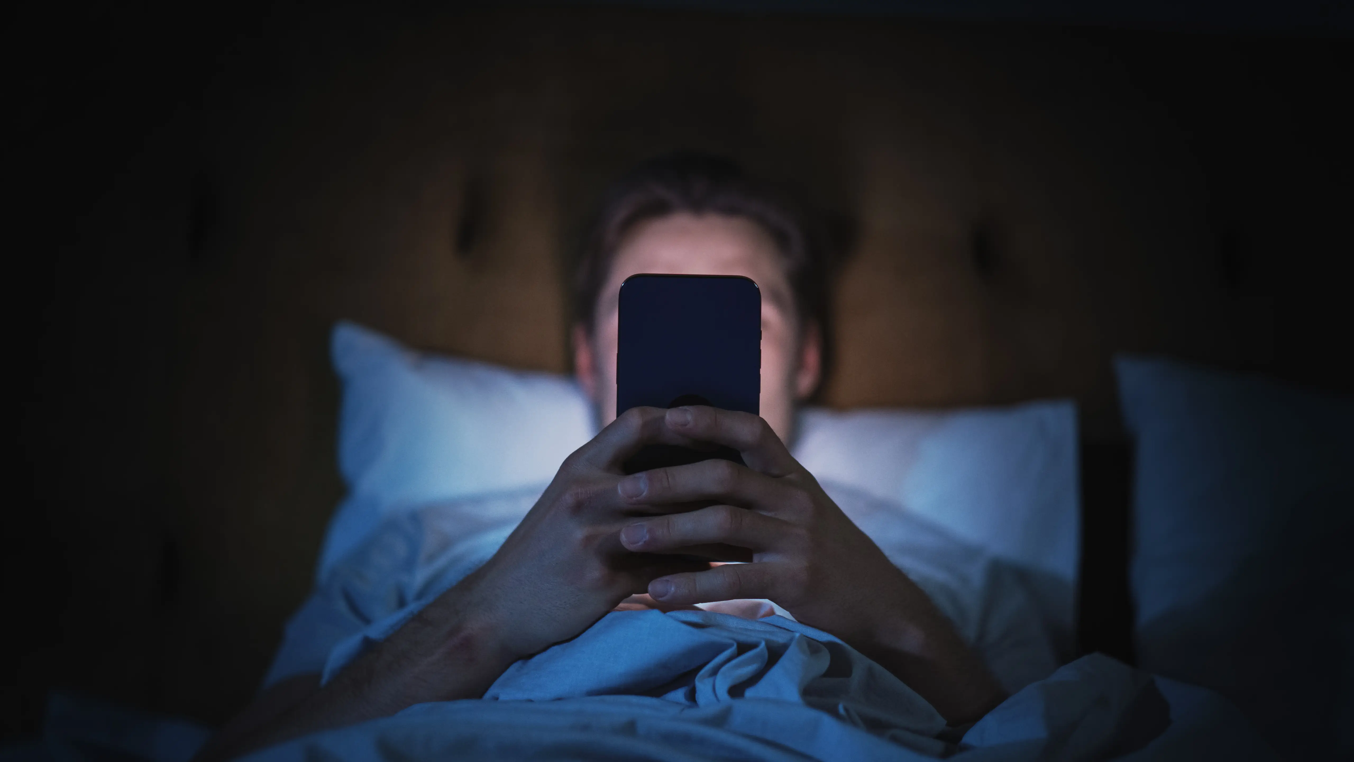Person lying in bed at night holds a smartphone illuminated by blue screen light.