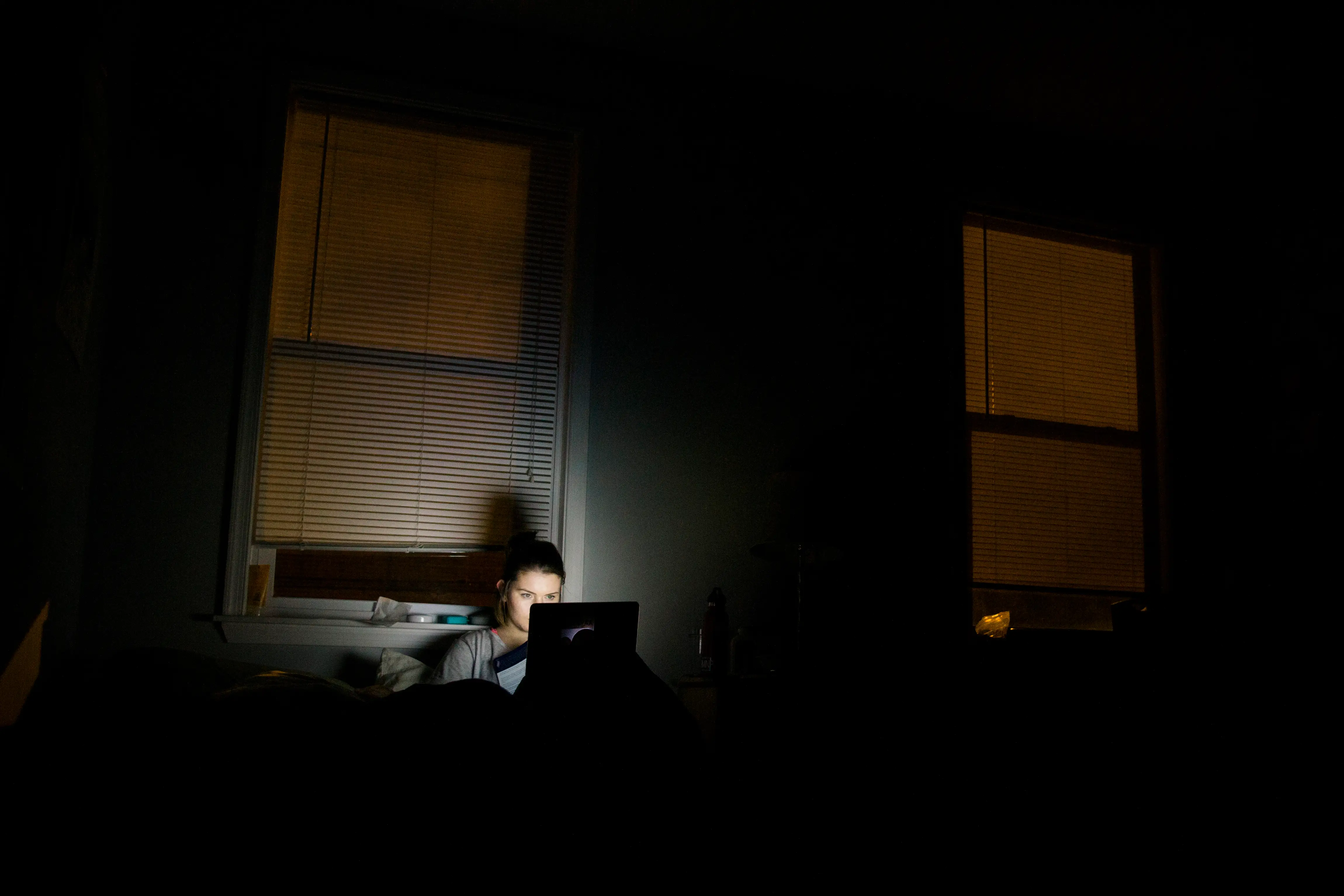 Person works on a laptop in a dark room with lit windows and closed blinds.