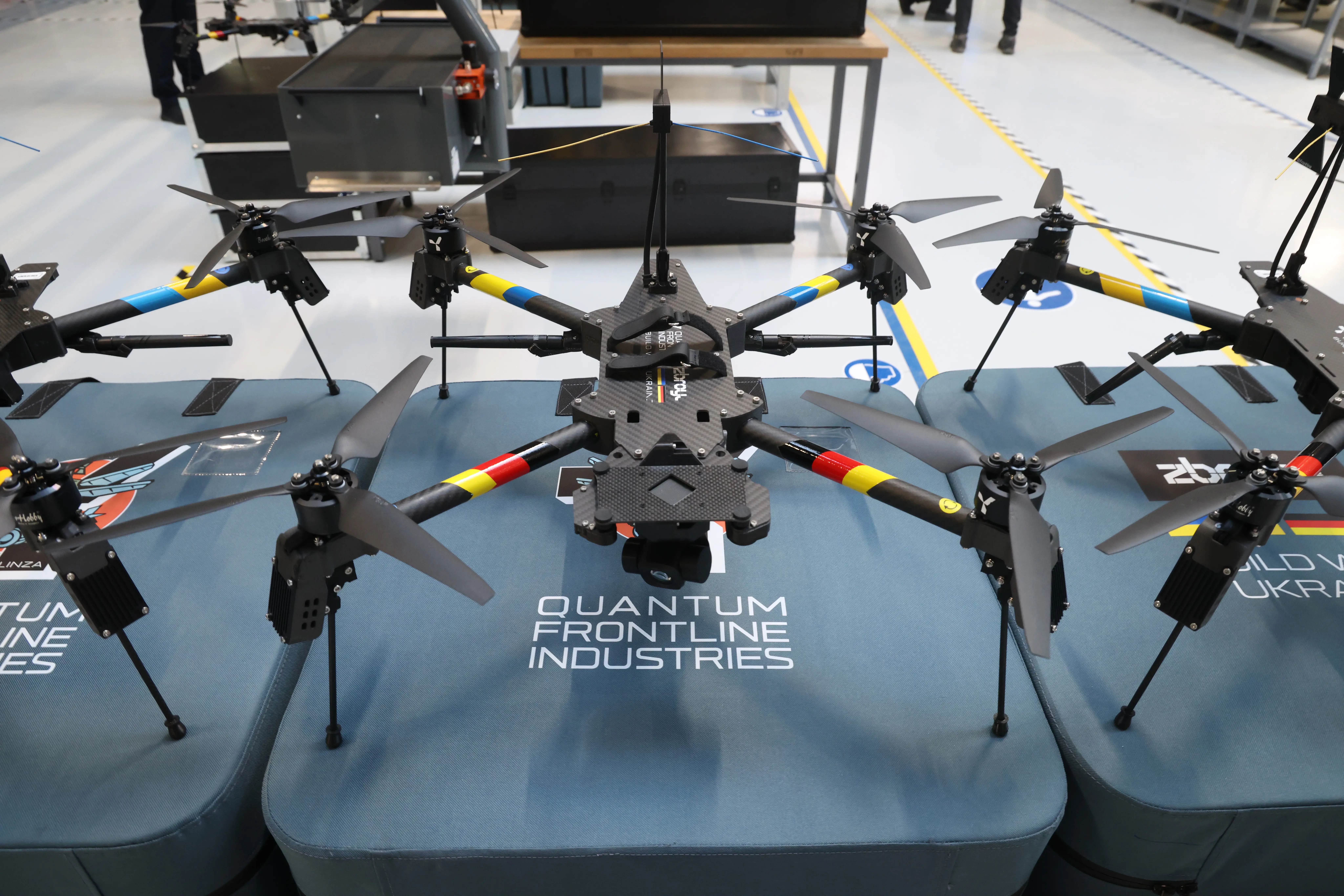 Multirotor drones are displayed on platforms labeled QUANTUM FRONTLINE INDUSTRIES in a clean industrial exhibition space.