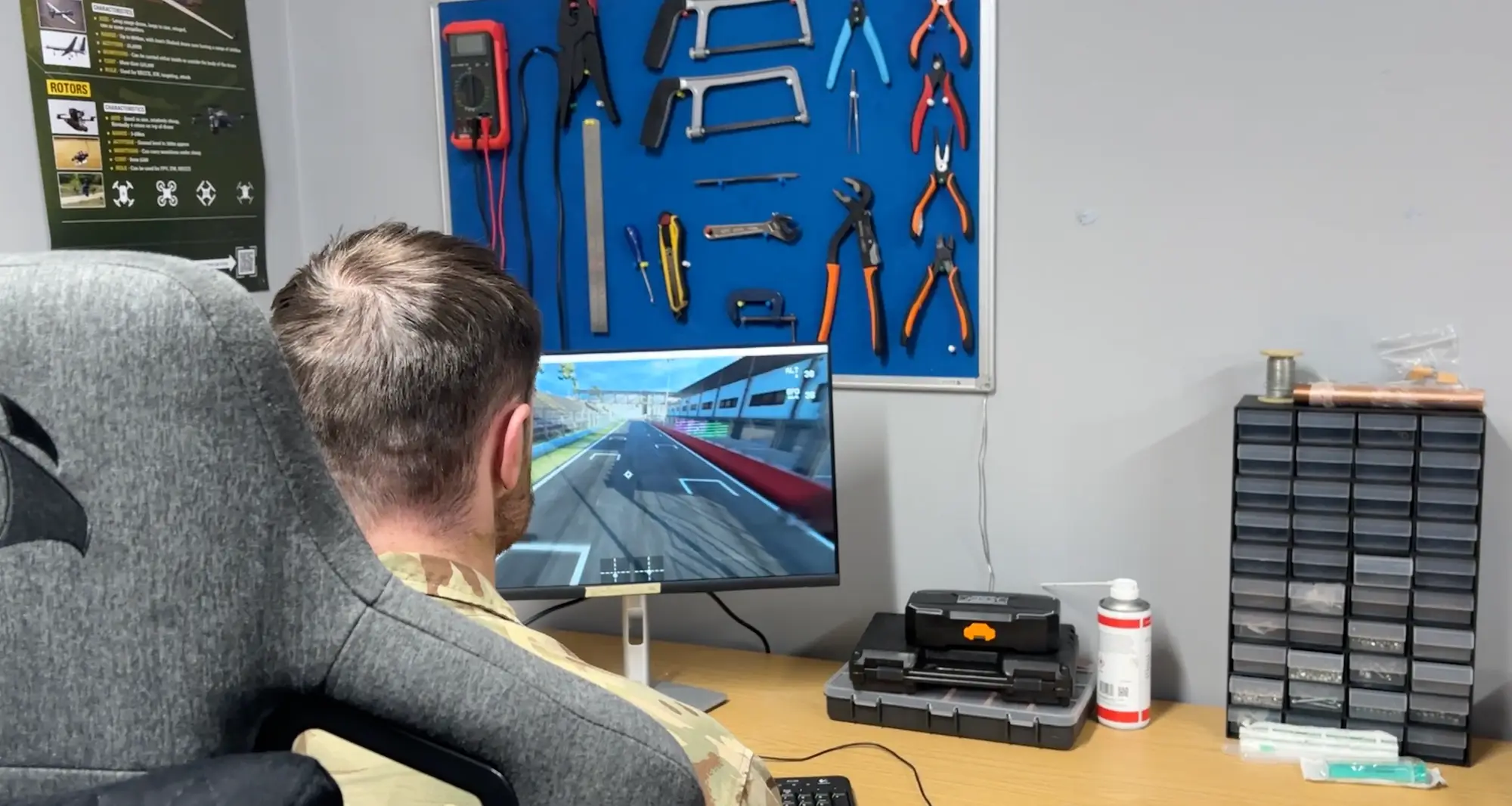 Person in camouflage clothing sits at a desk playing a racing simulator beside mounted tools and storage drawers.