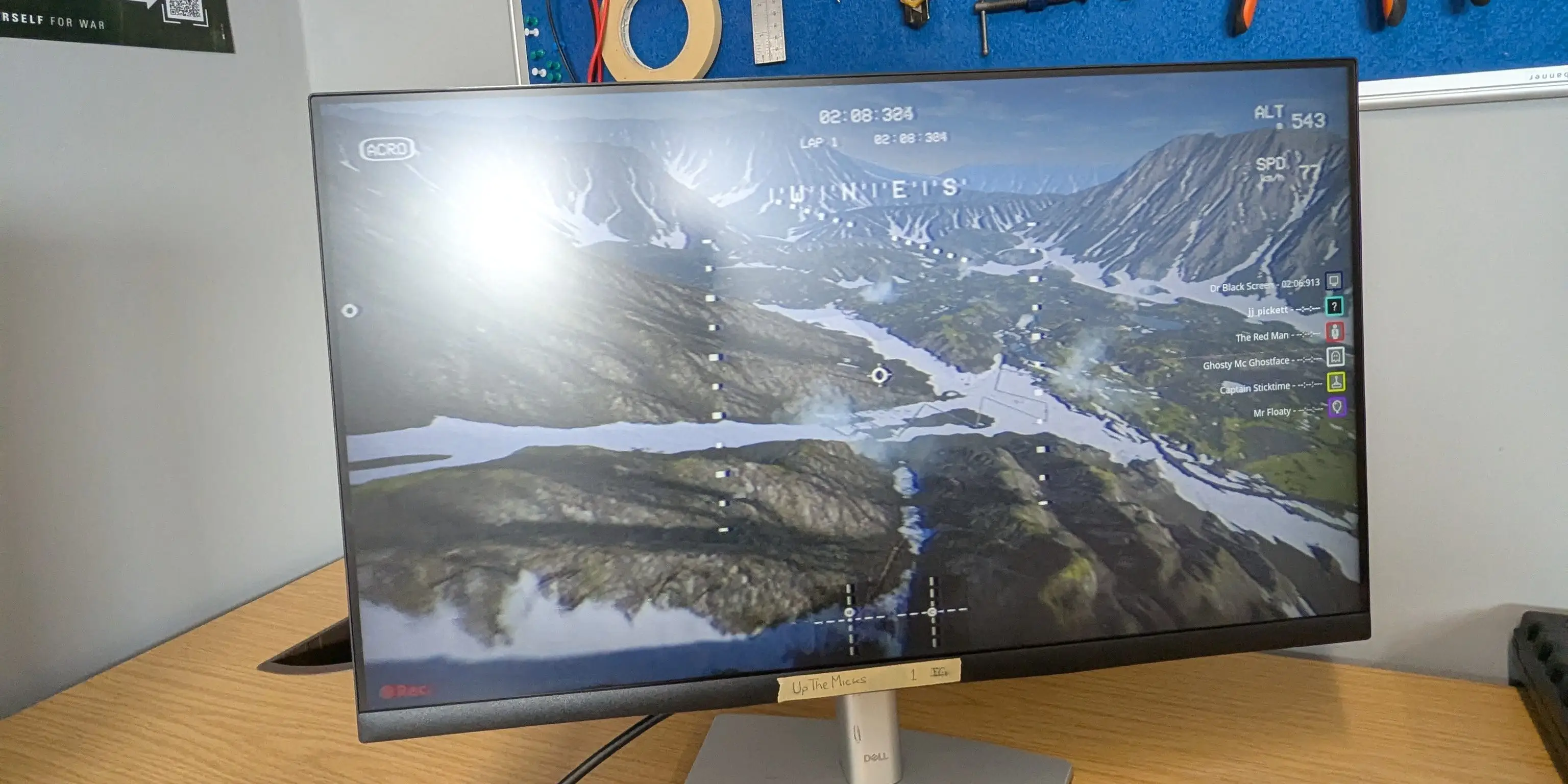 Computer monitor on a desk shows a mountain landscape simulation beneath a pegboard of hand tools.