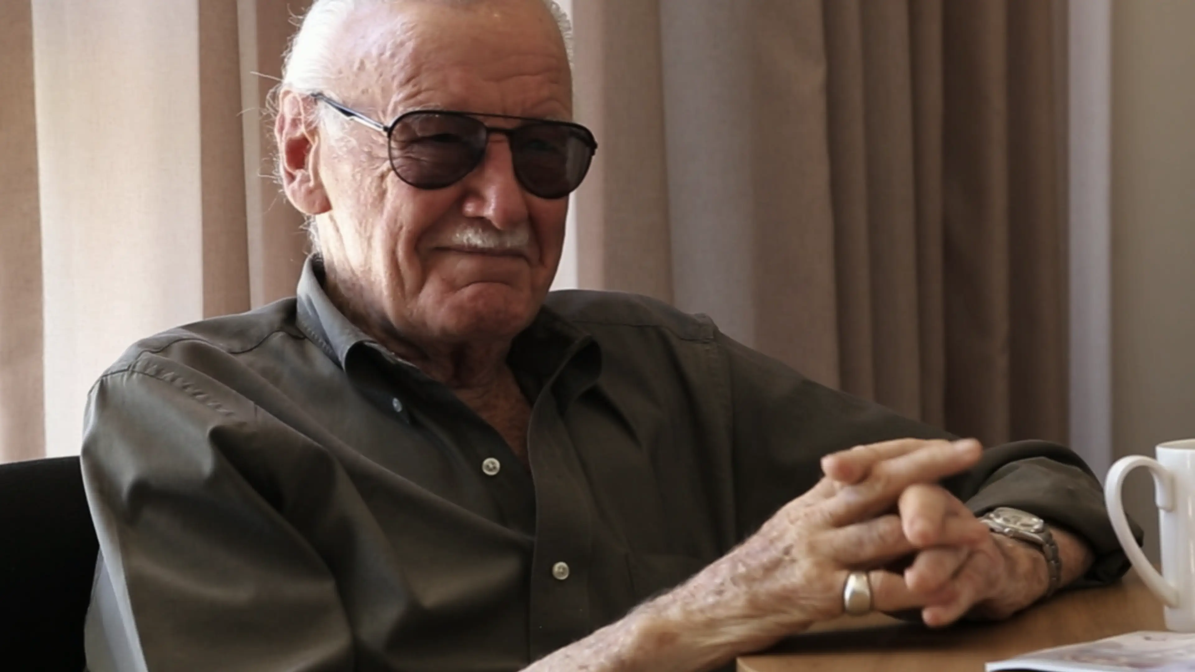 Stan Lee smiling hands folded on top of a table