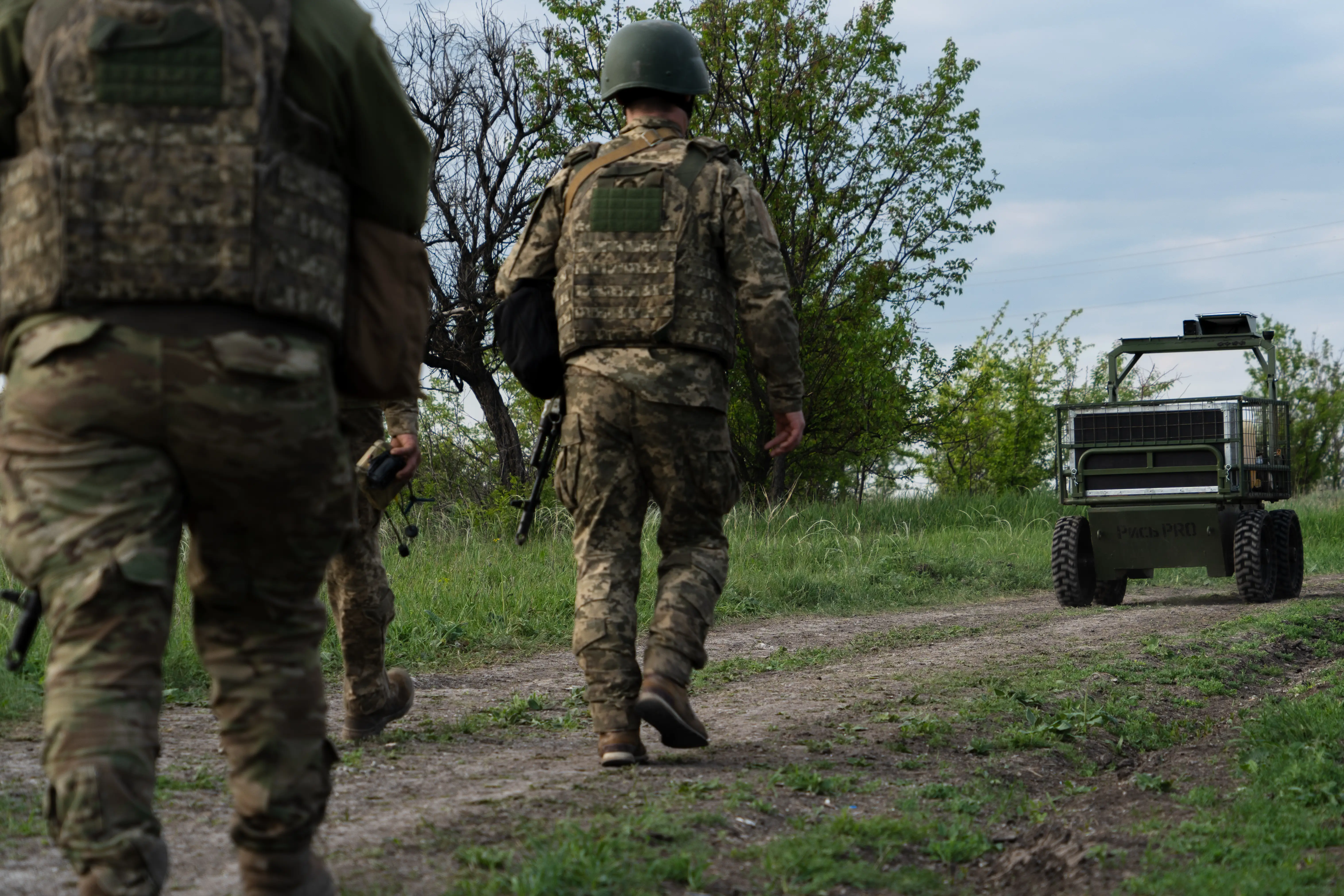 Ukrainian servicemen in full gear with rifles walk along a dirt path next to an unmanned ground vehicle on May 12, 2026, in Donetsk Oblast, Ukraine.