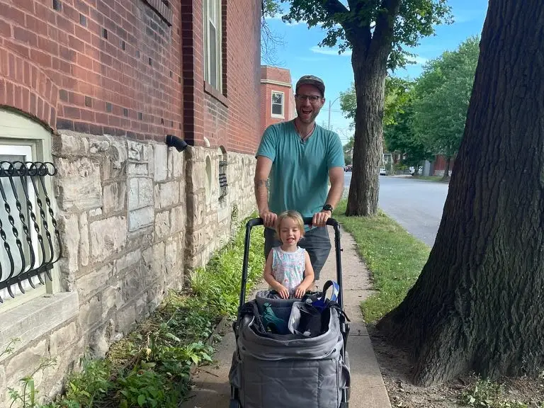 A smiling person pushes a gray stroller with a seated passenger along a tree-lined sidewalk beside brick buildings.