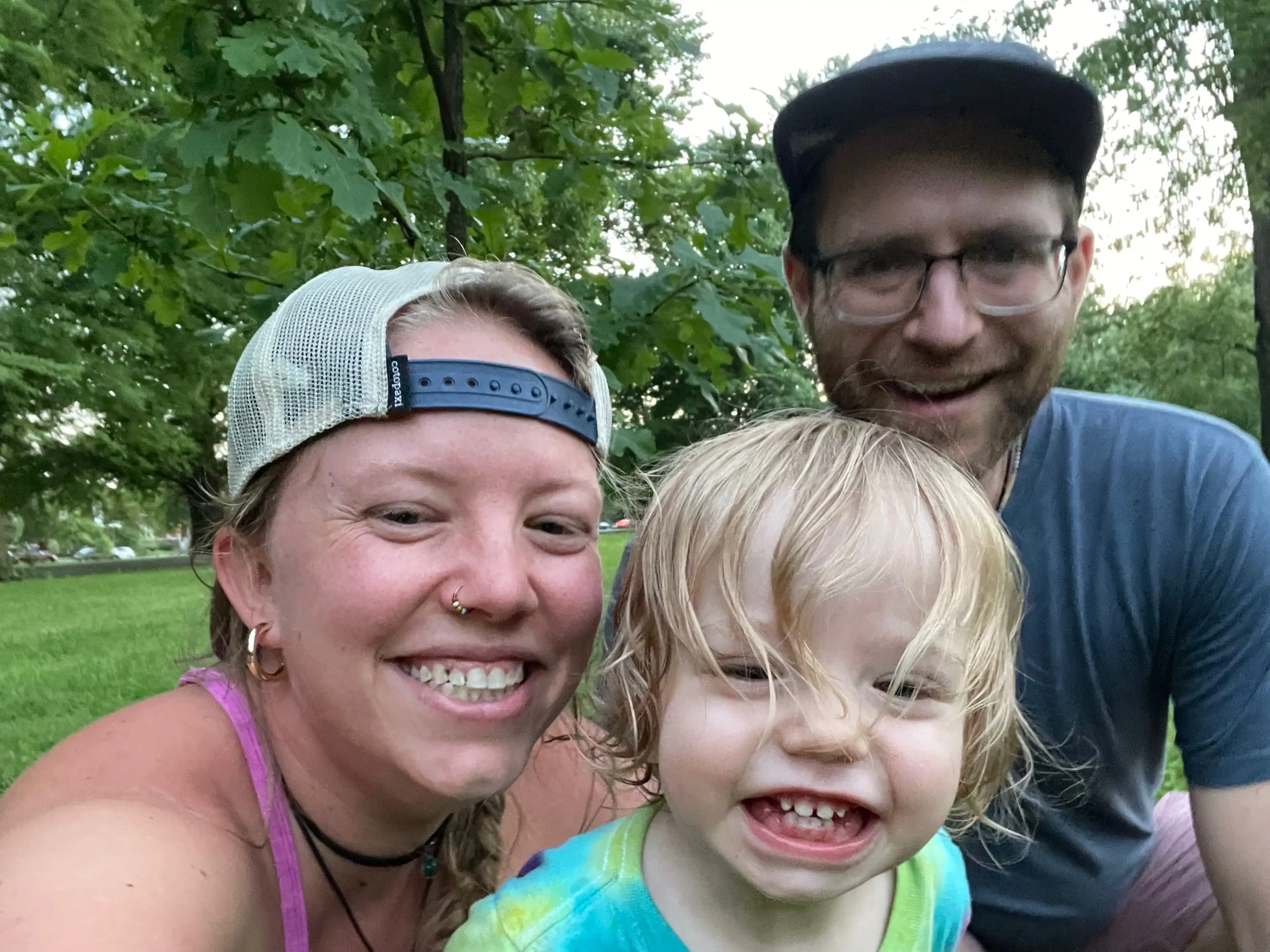 The writer, his spouse, and their kid posing while sitting in the grass on a sunny day.