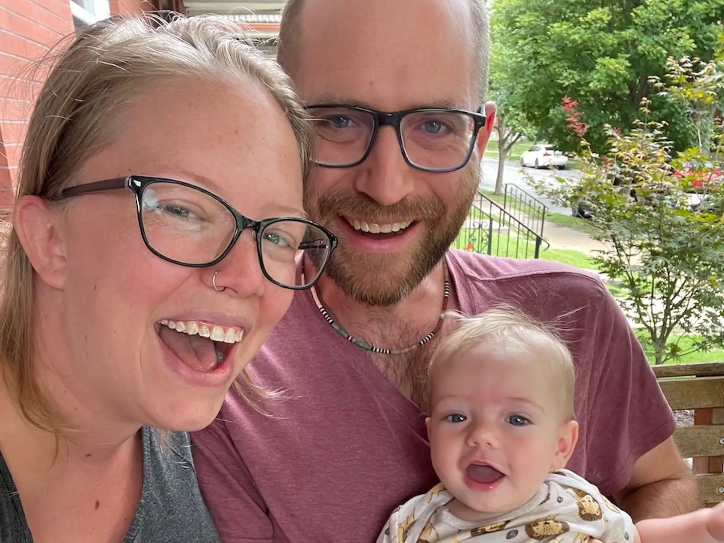 Three smiling people, including a child, sit close together on a porch with trees and a street behind them.