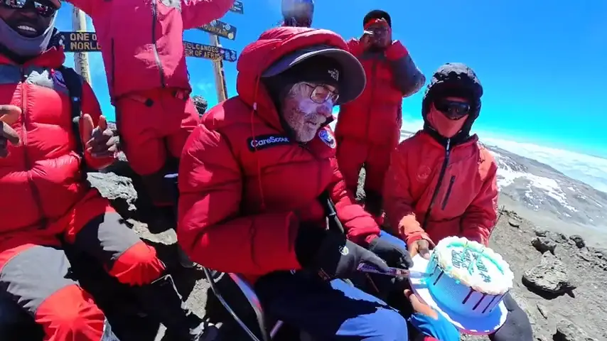 An older man cutting a cake at the summit of Kilimanjaro.