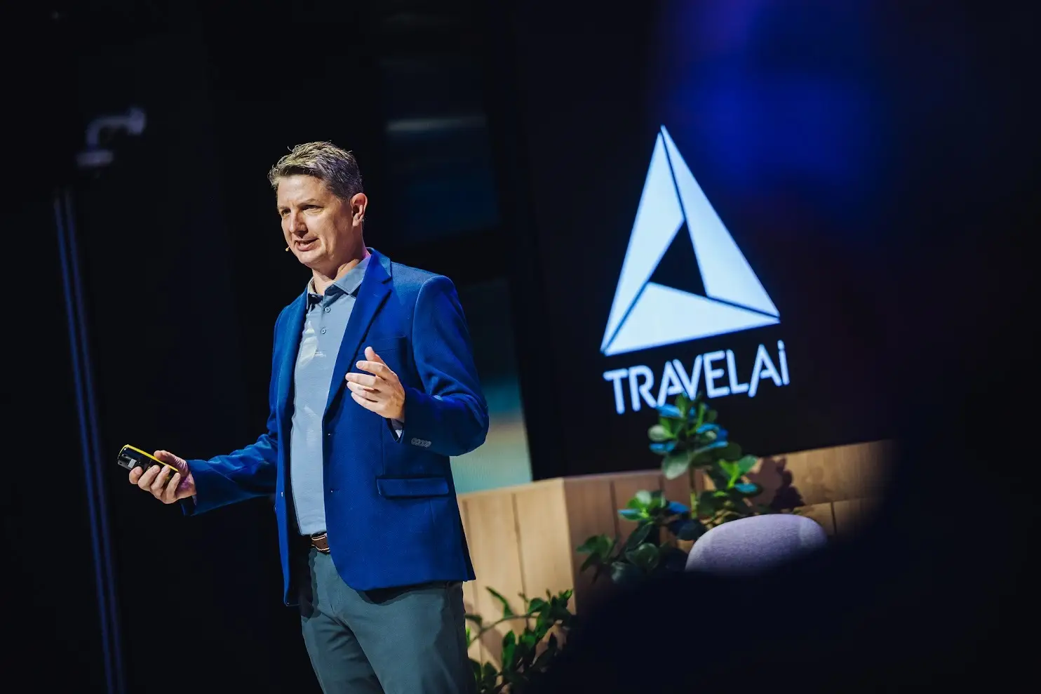 Presenter holding a clicker speaks onstage beside a large illuminated TRAVEL.Ai logo.