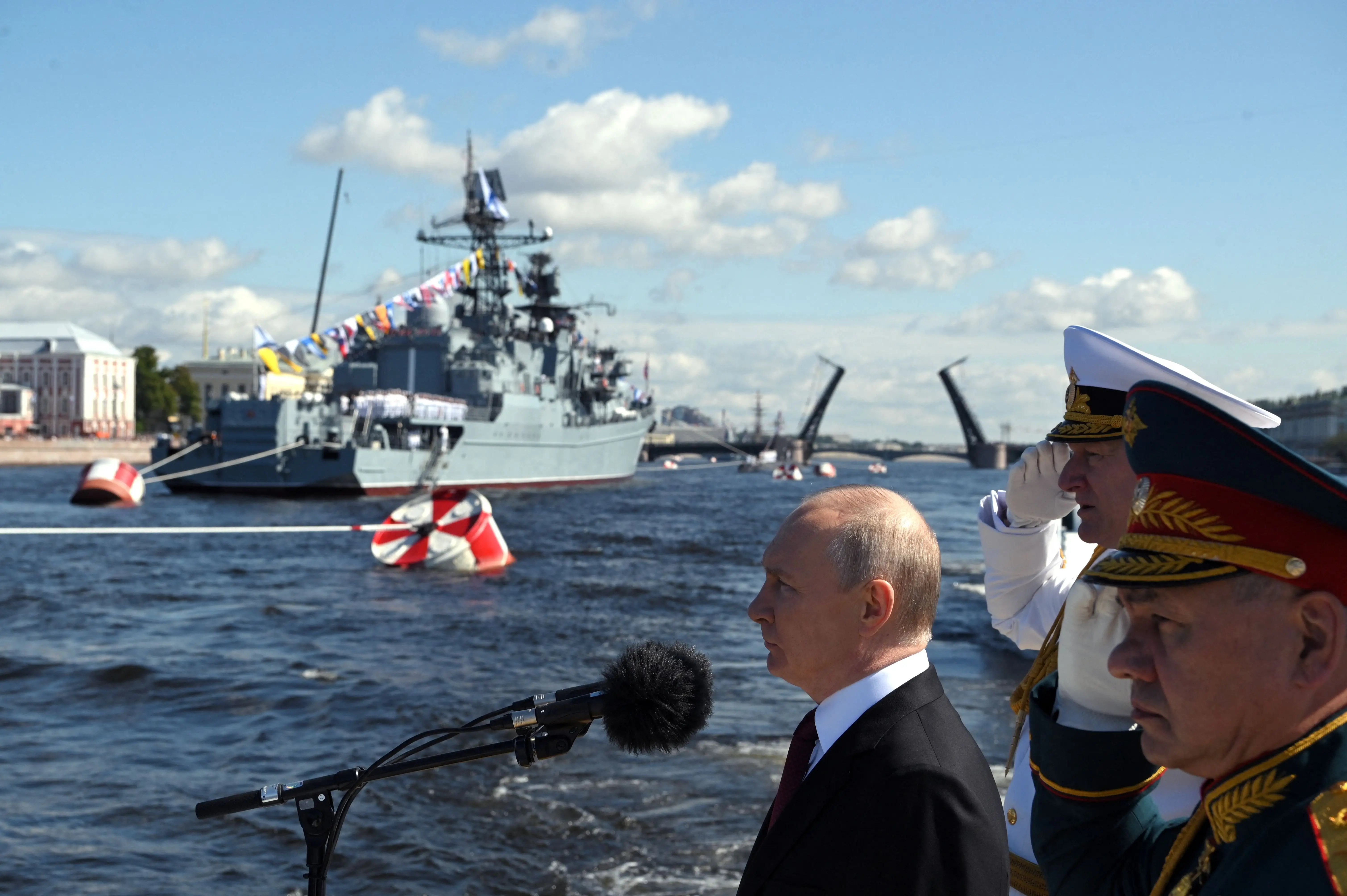Putin stands in front of a microphone with a warship in the background.