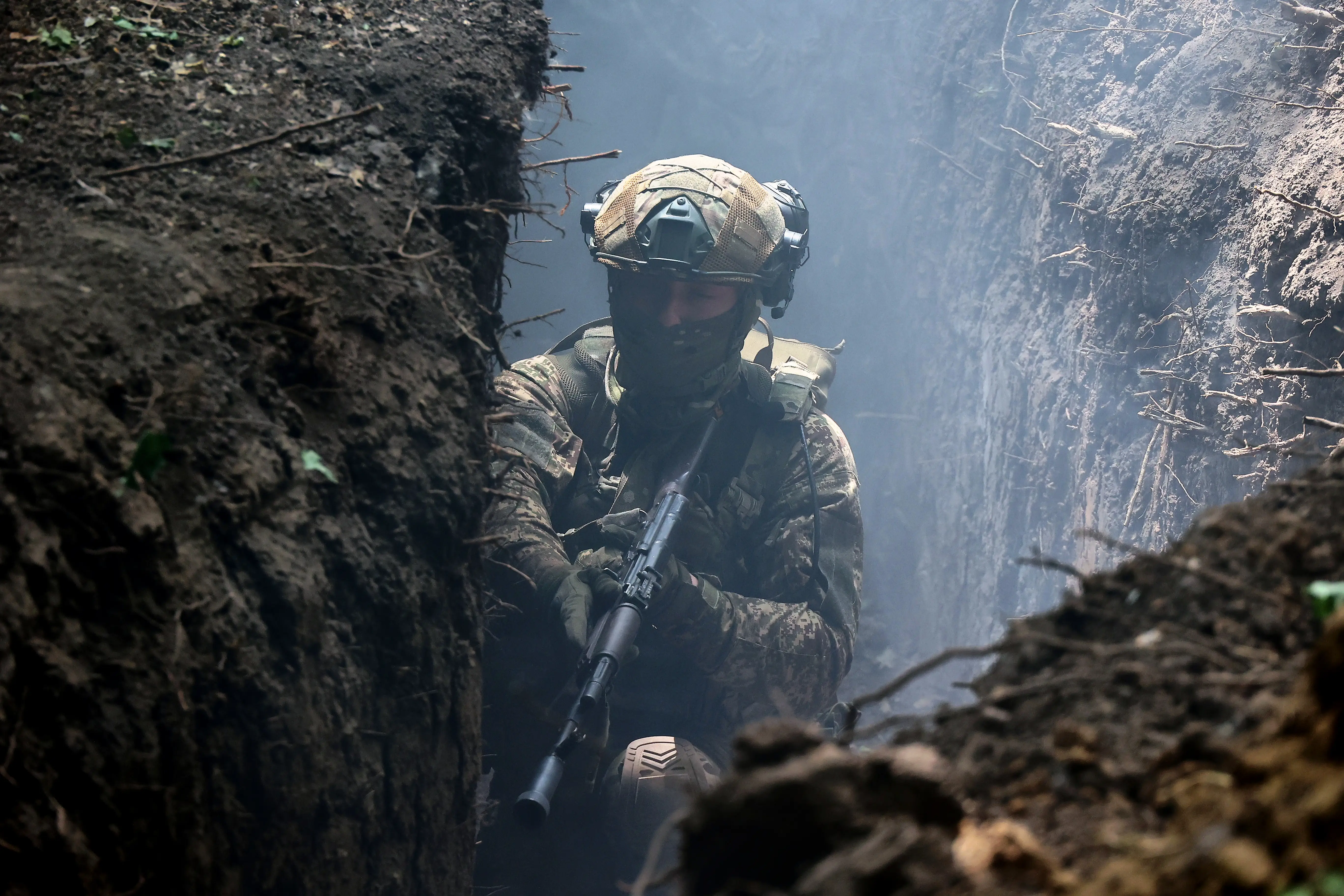 Camouflaged soldier holding a rifle moves through a narrow smoky earthen trench.