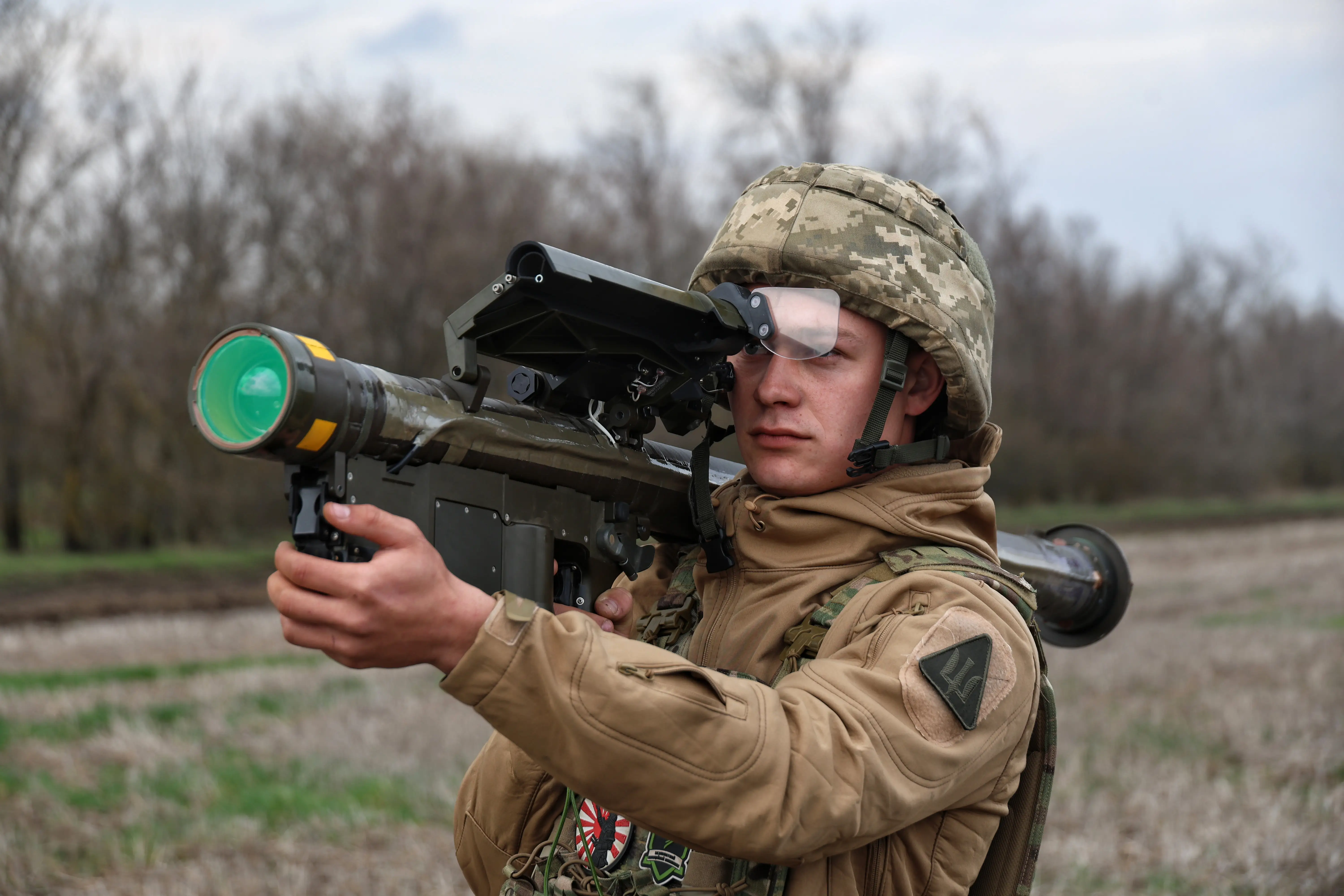 Person in camouflage helmet and tactical gear aims a shoulder-fired launcher in an open field.