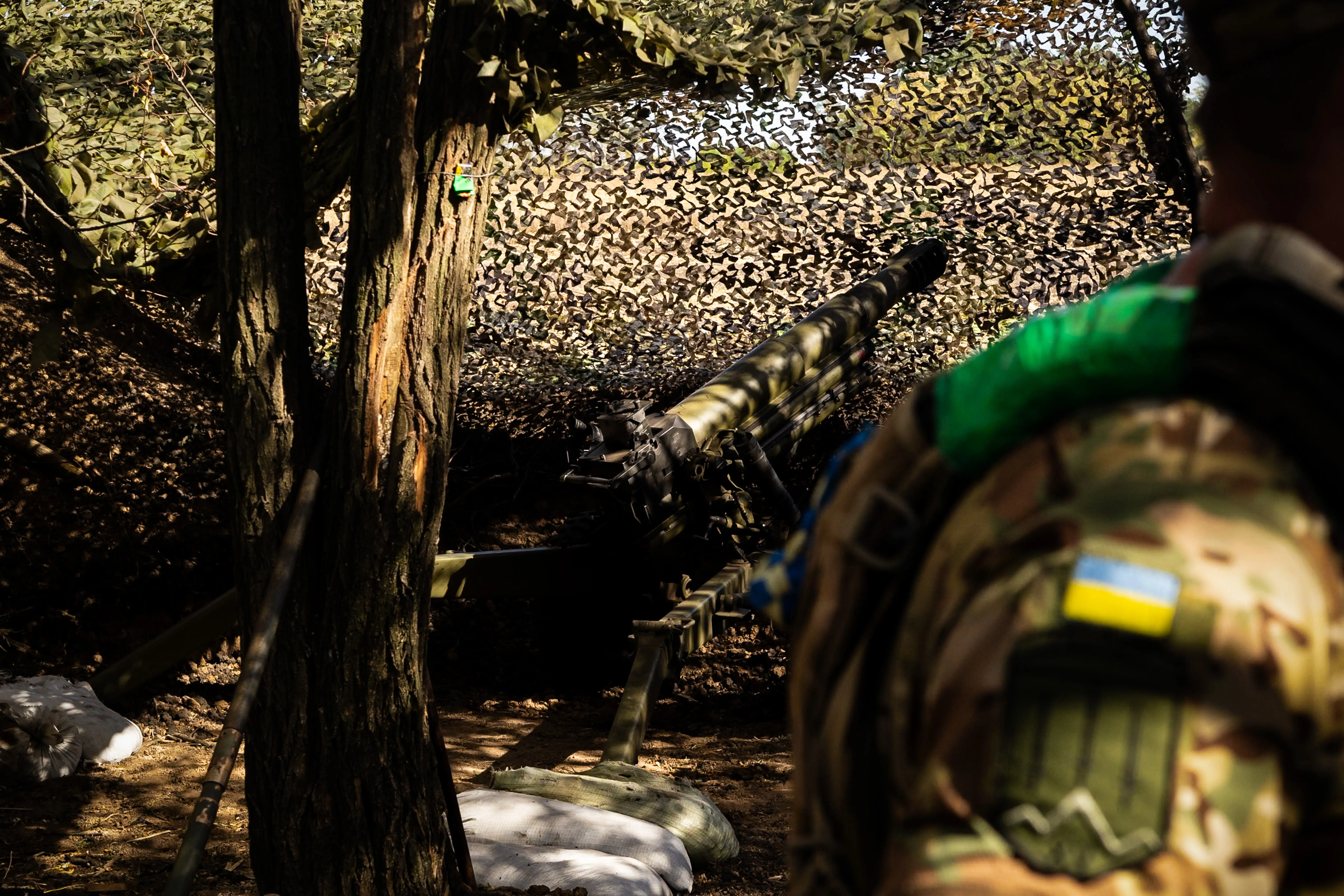 Camouflaged service member with a Ukrainian flag patch stands near artillery hidden under netting in a wooded area.