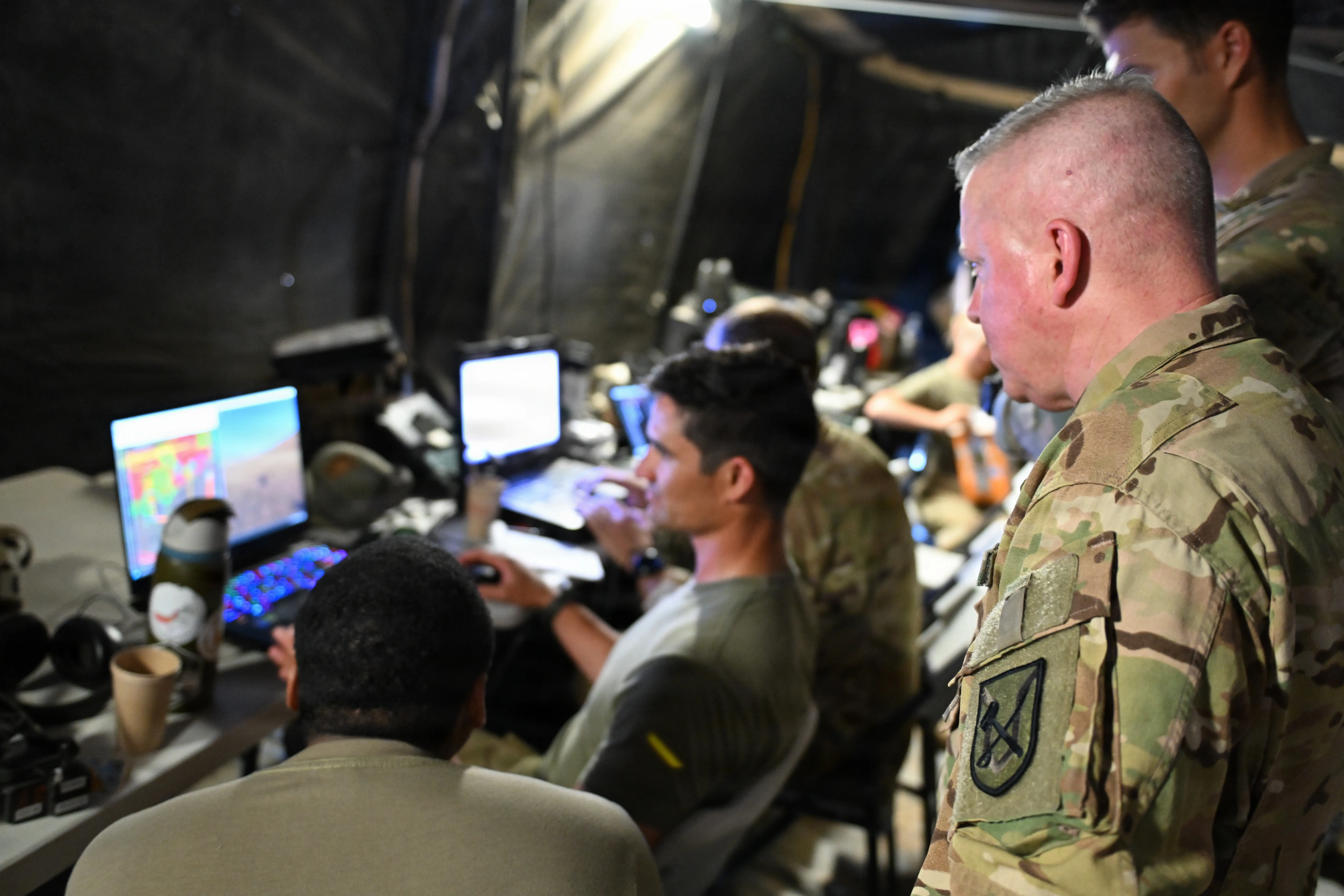 US Army soldiers stand and sit around laptops sitting on a table.