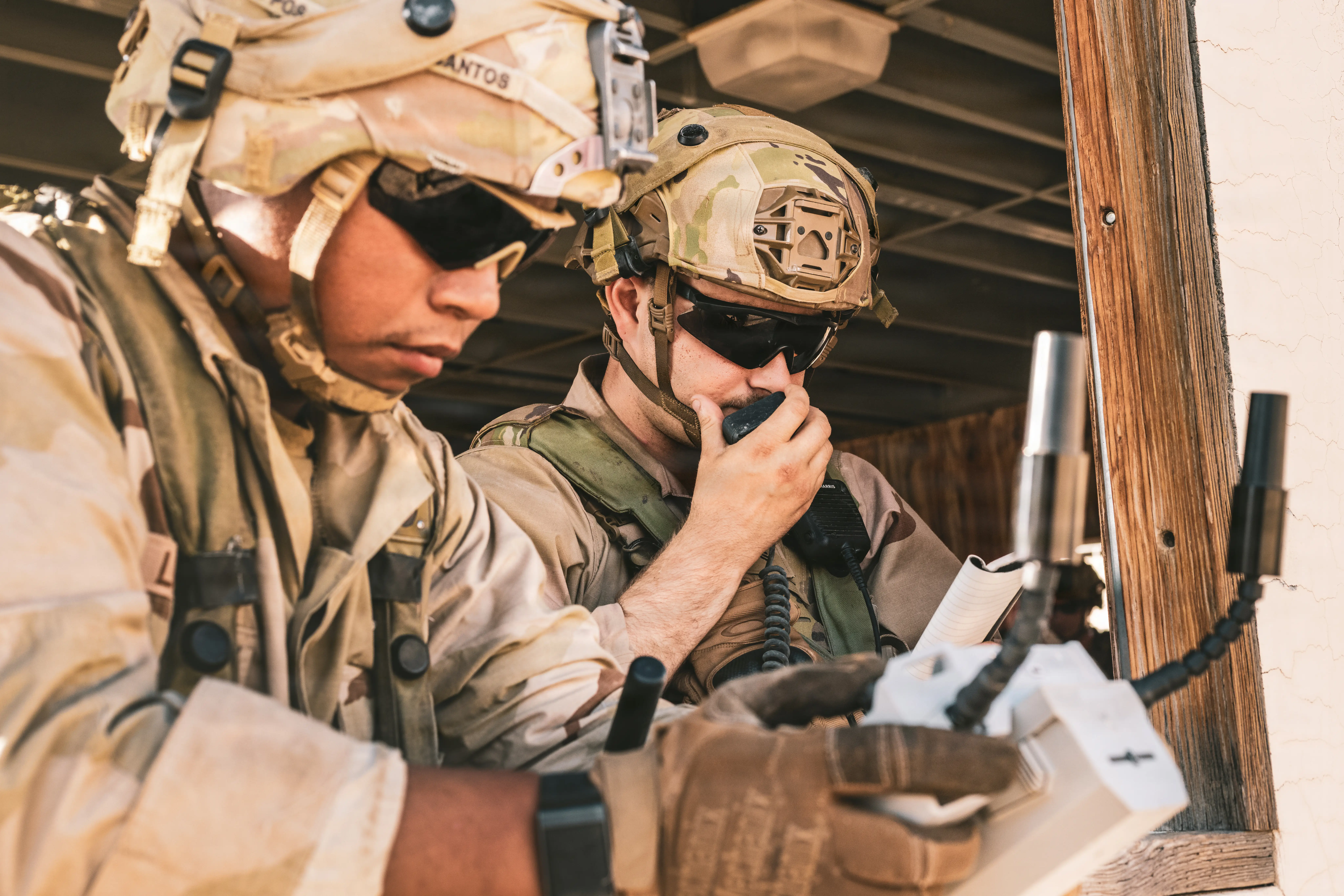 US Army soldiers stand in a window, one holding a radio to his mouth and the other looking at a monitor in his hands.