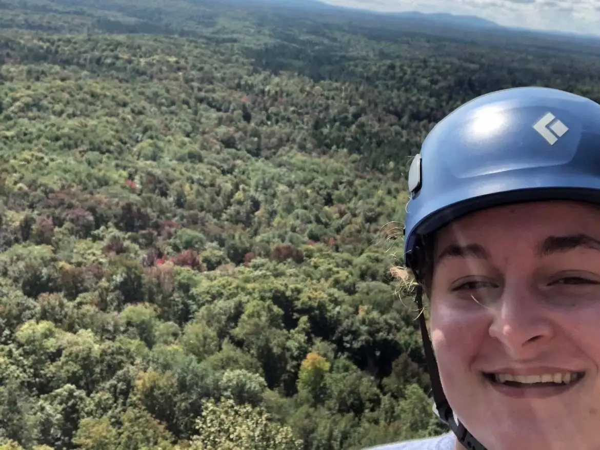 Smiling helmeted person takes a selfie above a broad forested landscape under a partly cloudy sky.