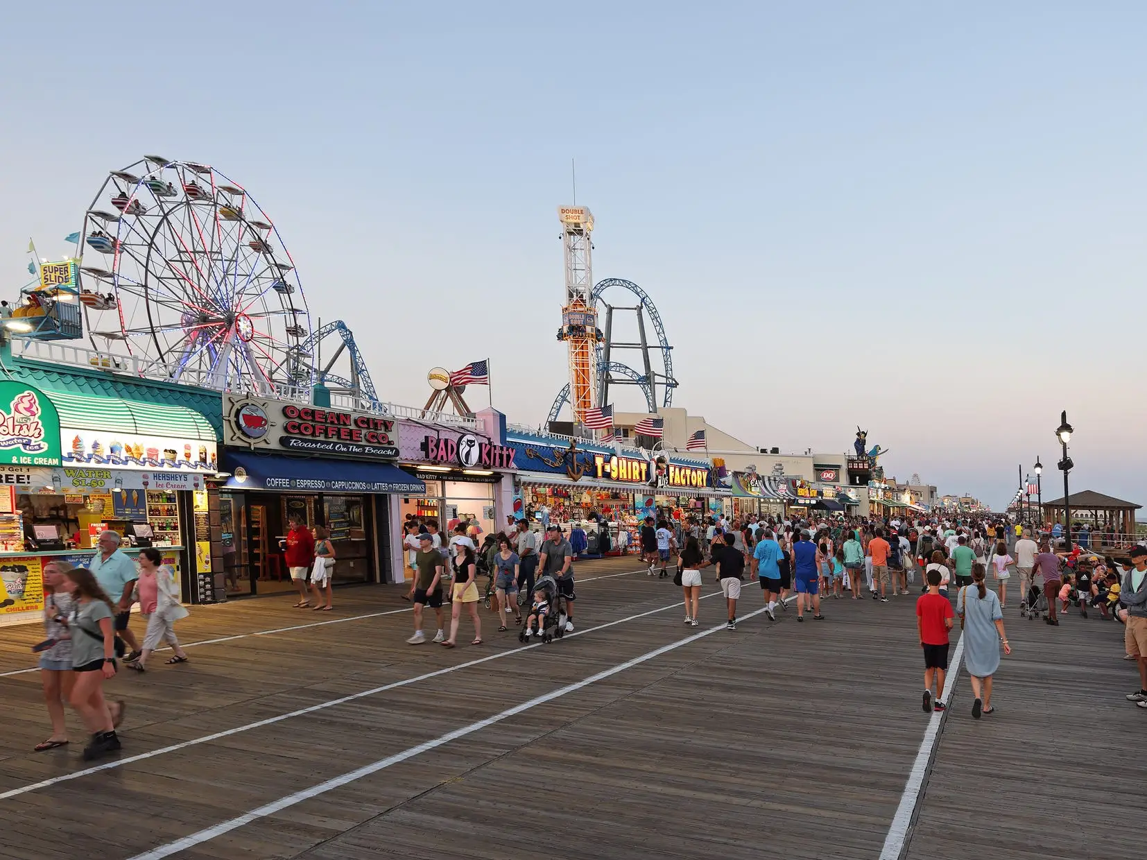People walking on a boardwalk lined with shops at dusk. There are rides in the background.