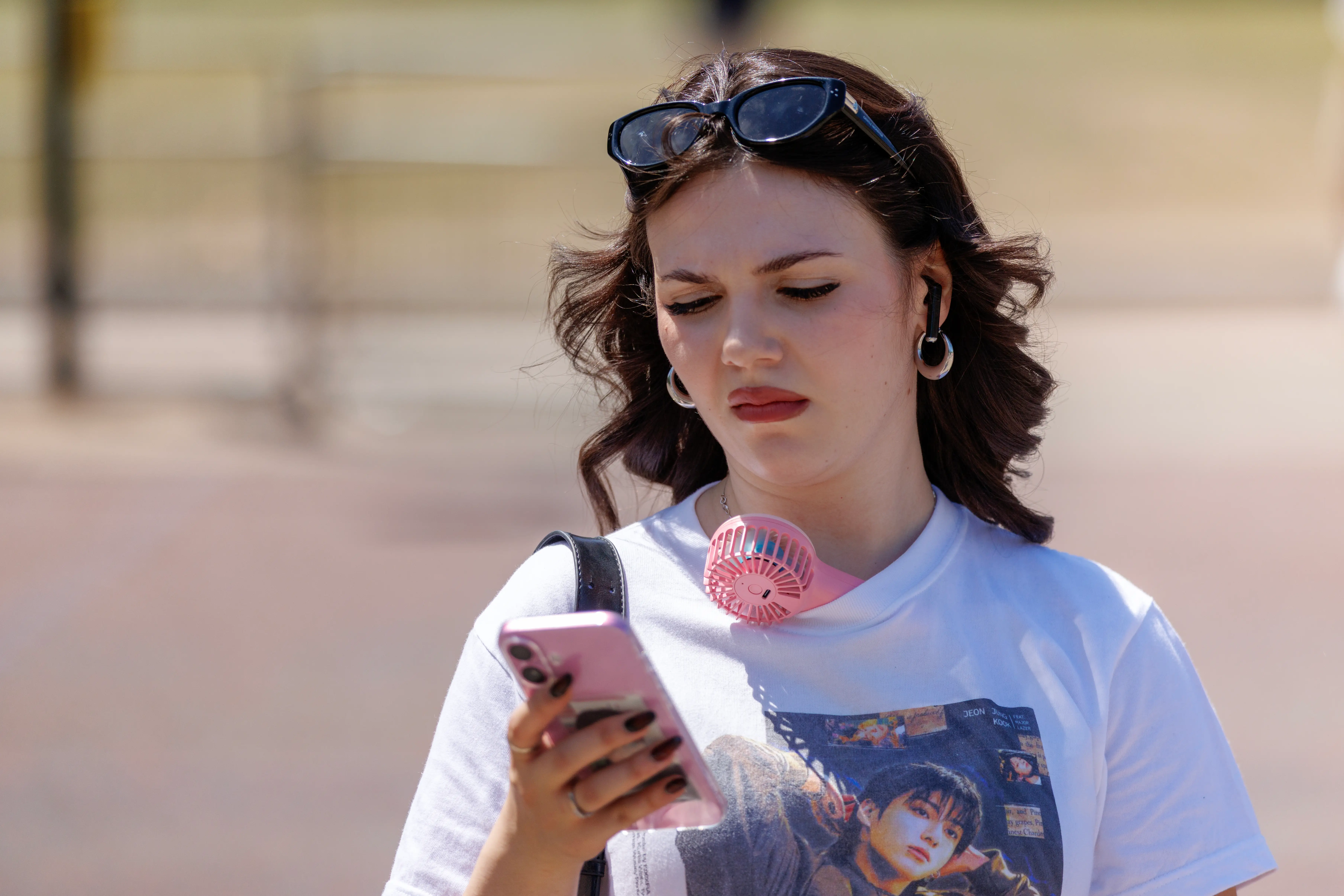 Person outdoors looks at a pink smartphone while wearing sunglasses on their head and a small pink neck fan.