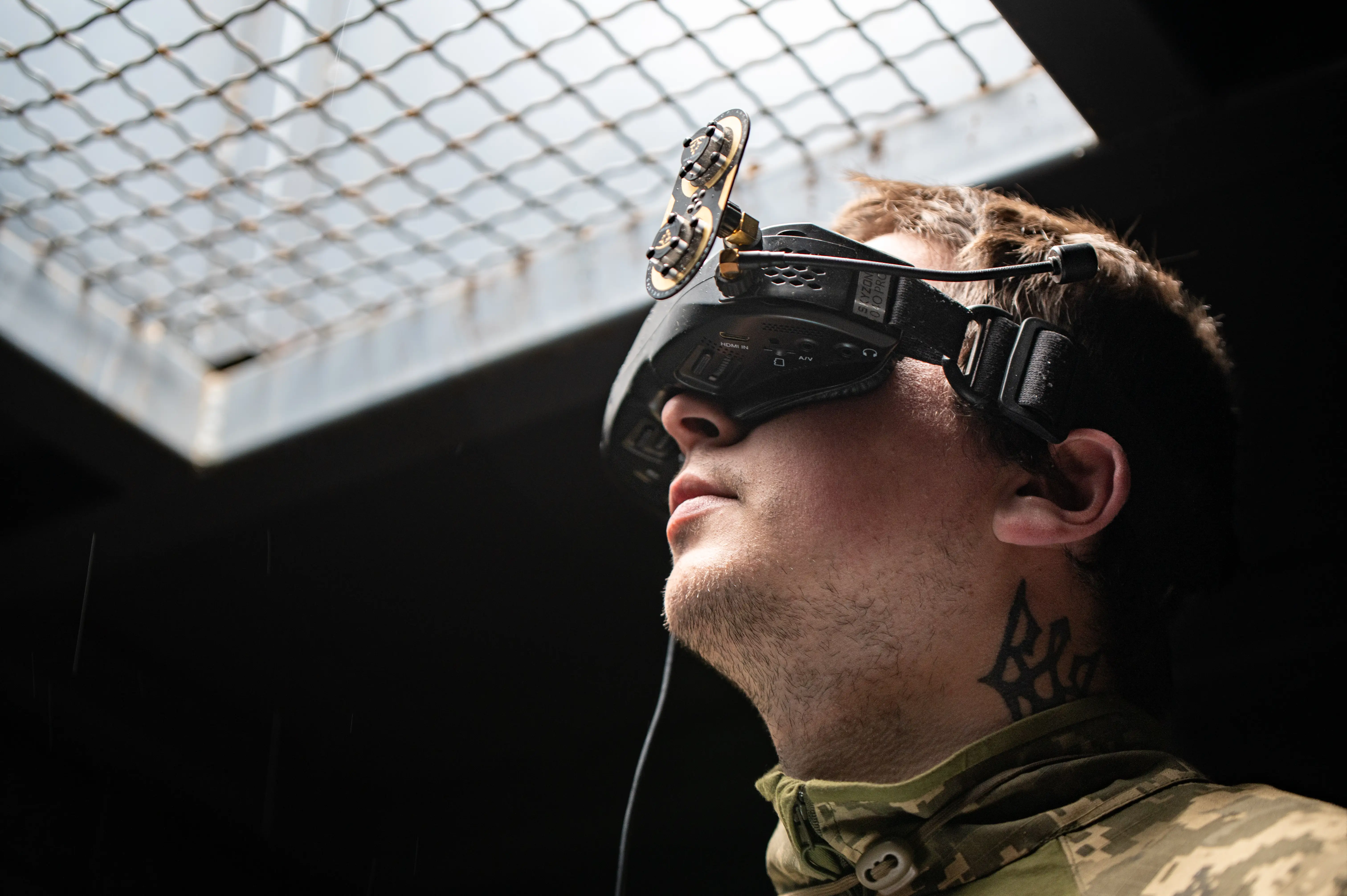 Person in camouflage clothing wears FPV goggles beneath a metal mesh opening with sunlight overhead.