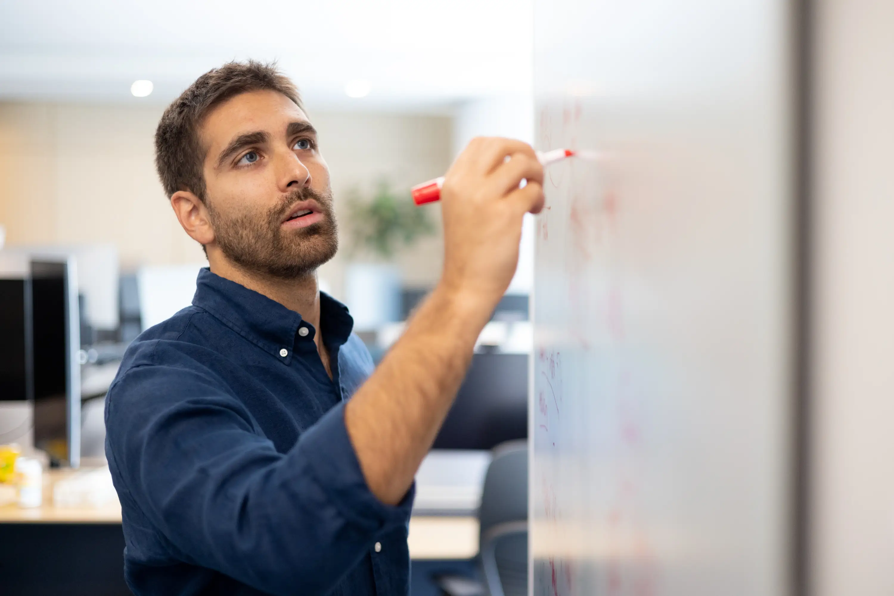 Person in a blue shirt writes with a red marker on a whiteboard in an office setting.