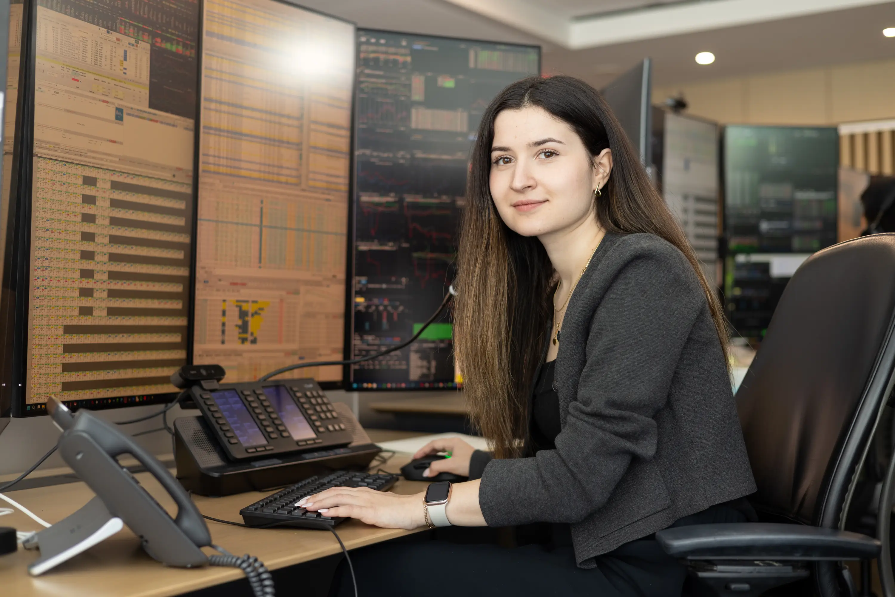 Person seated at a multi-monitor workstation with financial charts, data screens, phones, keyboard, and mouse.