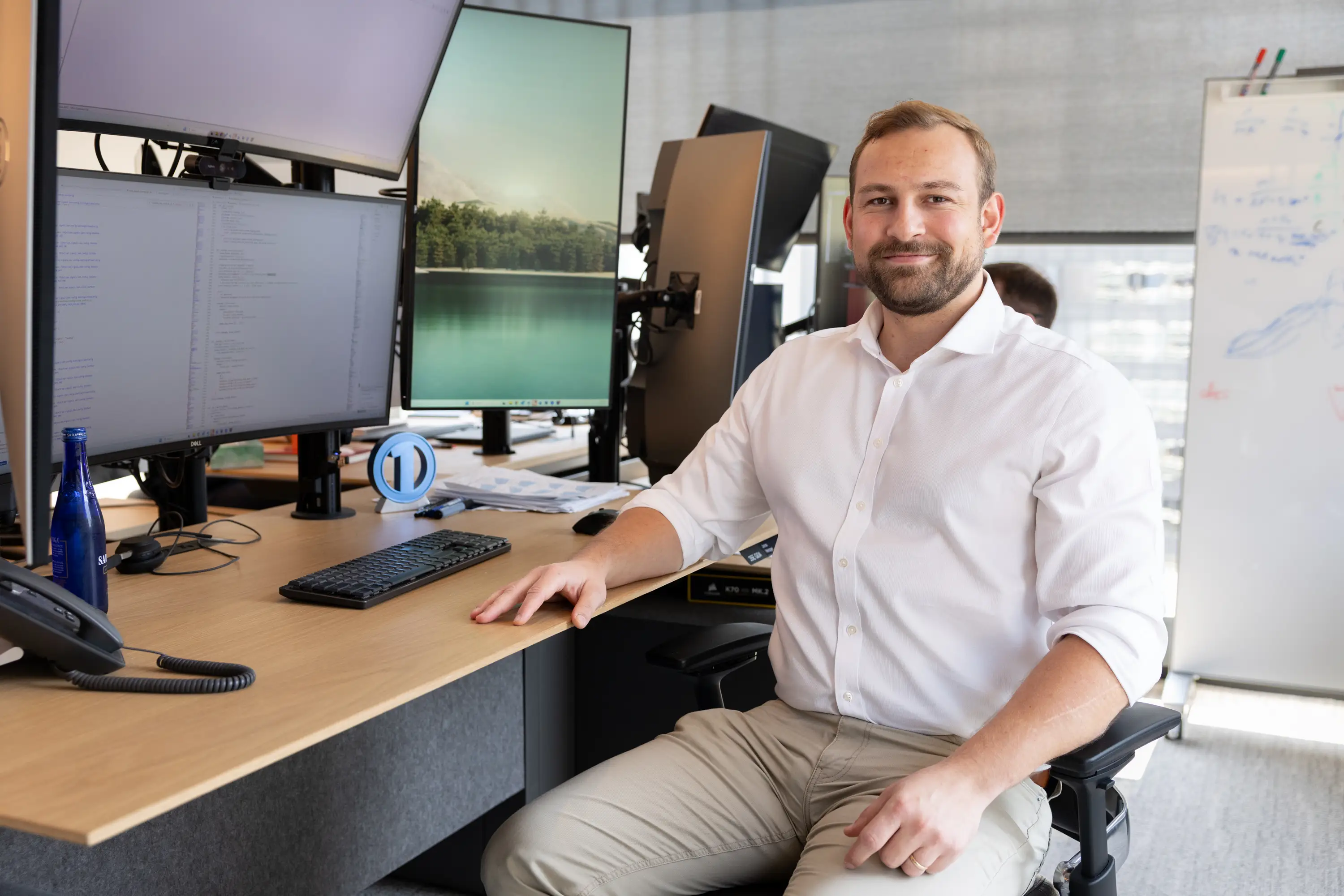 Office worker seated at a desk with multiple monitors, keyboard, phone, and a whiteboard in the background.
