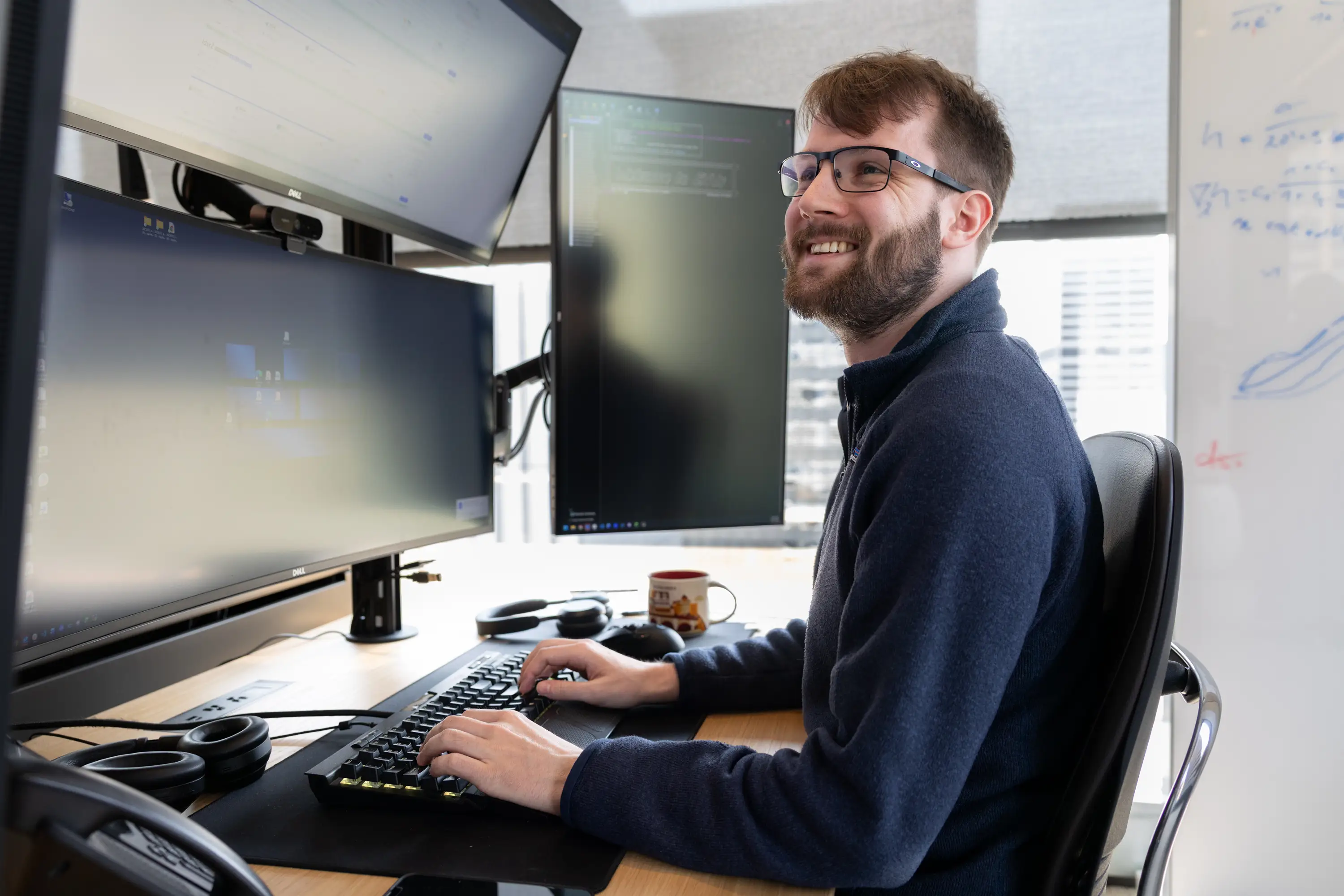 Person wearing glasses smiles while typing at a desk with multiple monitors and a whiteboard nearby.