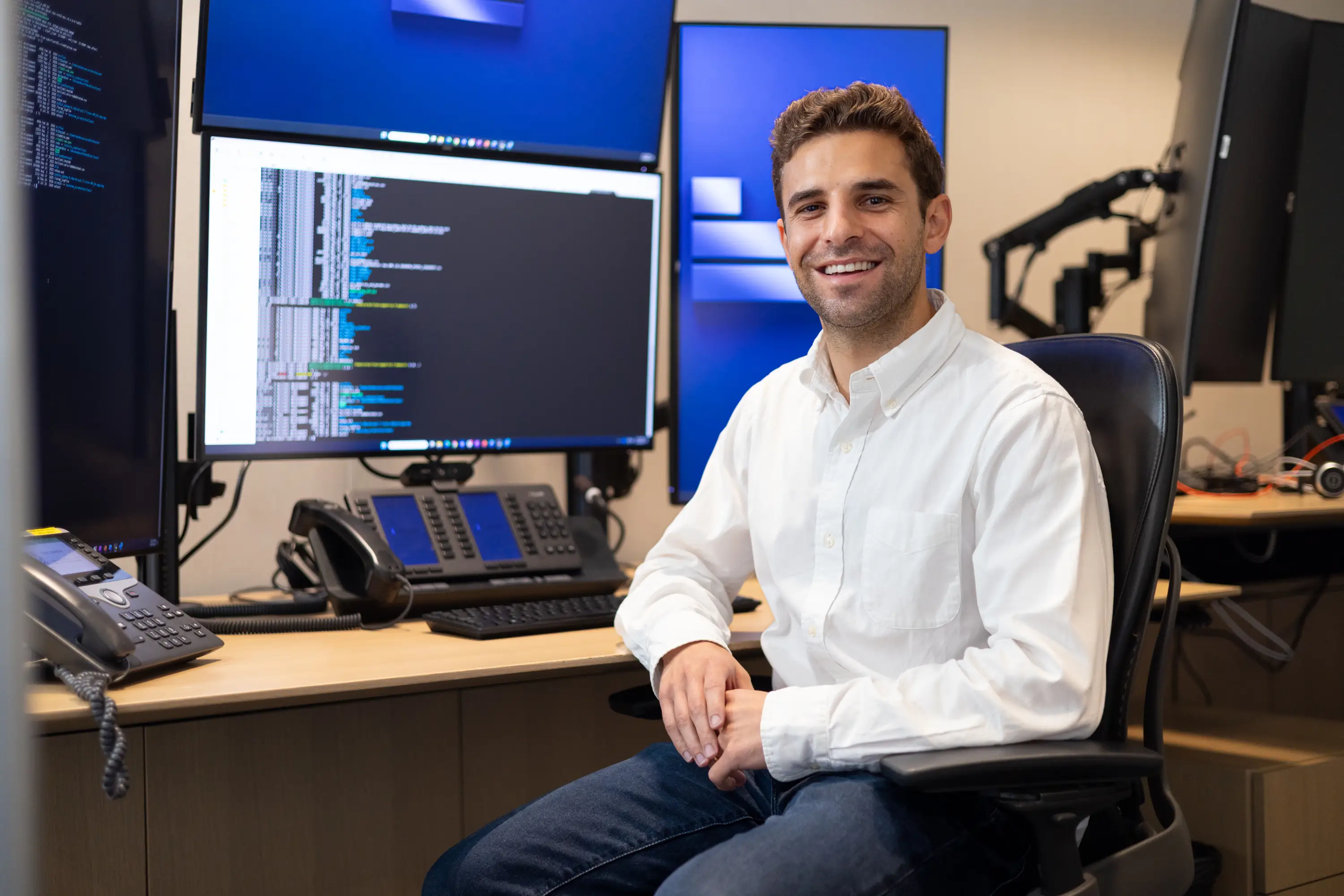 Smiling person seated at an office workstation with multiple monitors displaying software code.
