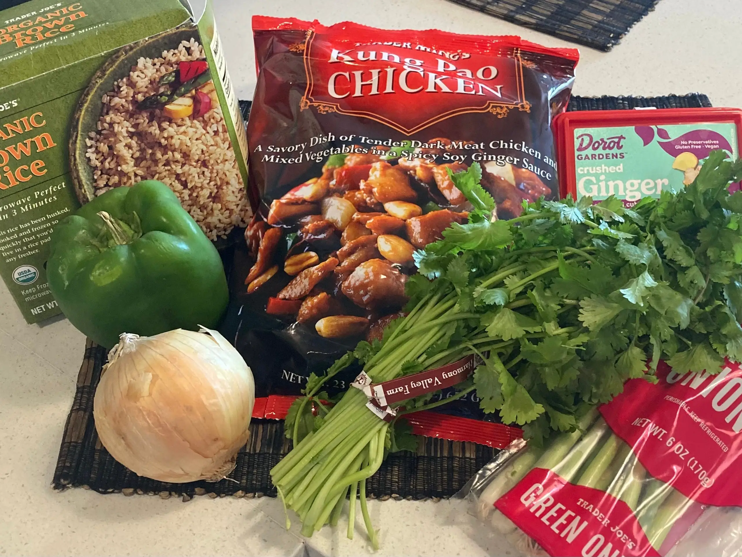 A bag of Trader Joe's kung pao chicken lying on a table next to some cilantro, a bell pepper, green onions, ginger, and a box of Trader Joe's organic brown rice.
