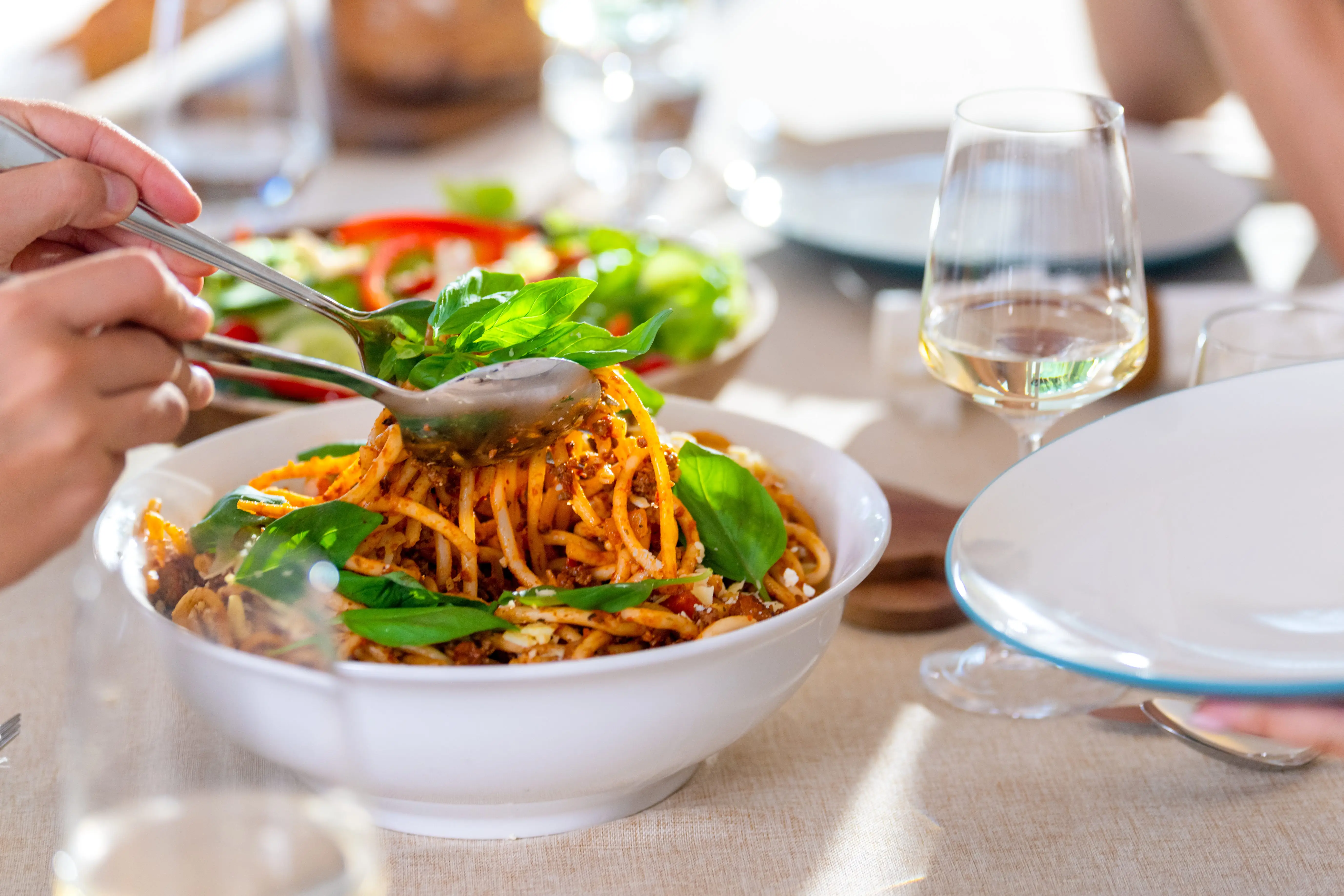 Hands serve spaghetti with tomato sauce and basil from a white bowl on a sunlit dining table.