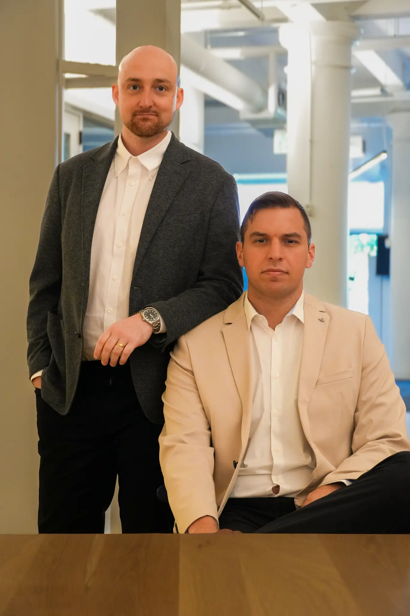 Two adults in business attire pose in a modern office beside a wooden table.