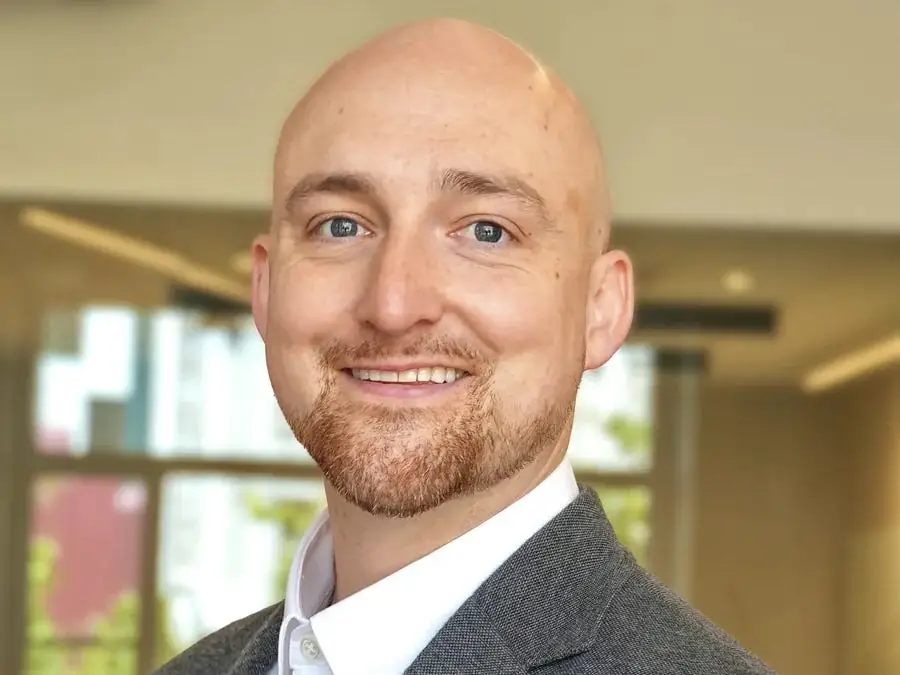 Smiling person in a gray blazer and white shirt poses for a professional indoor portrait.