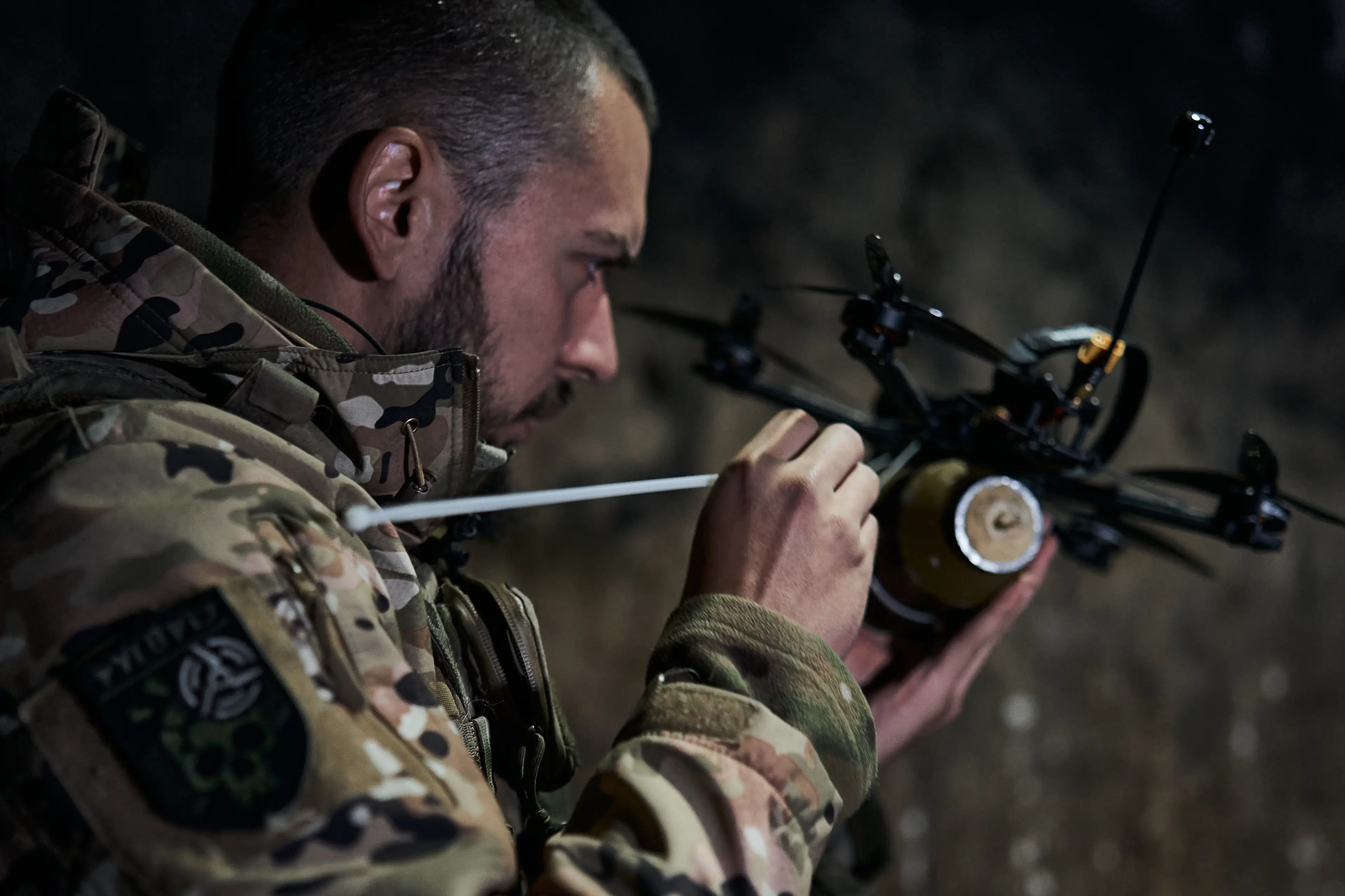 A camouflaged uniformed person adjusts a small quadcopter drone in a dim outdoor setting.