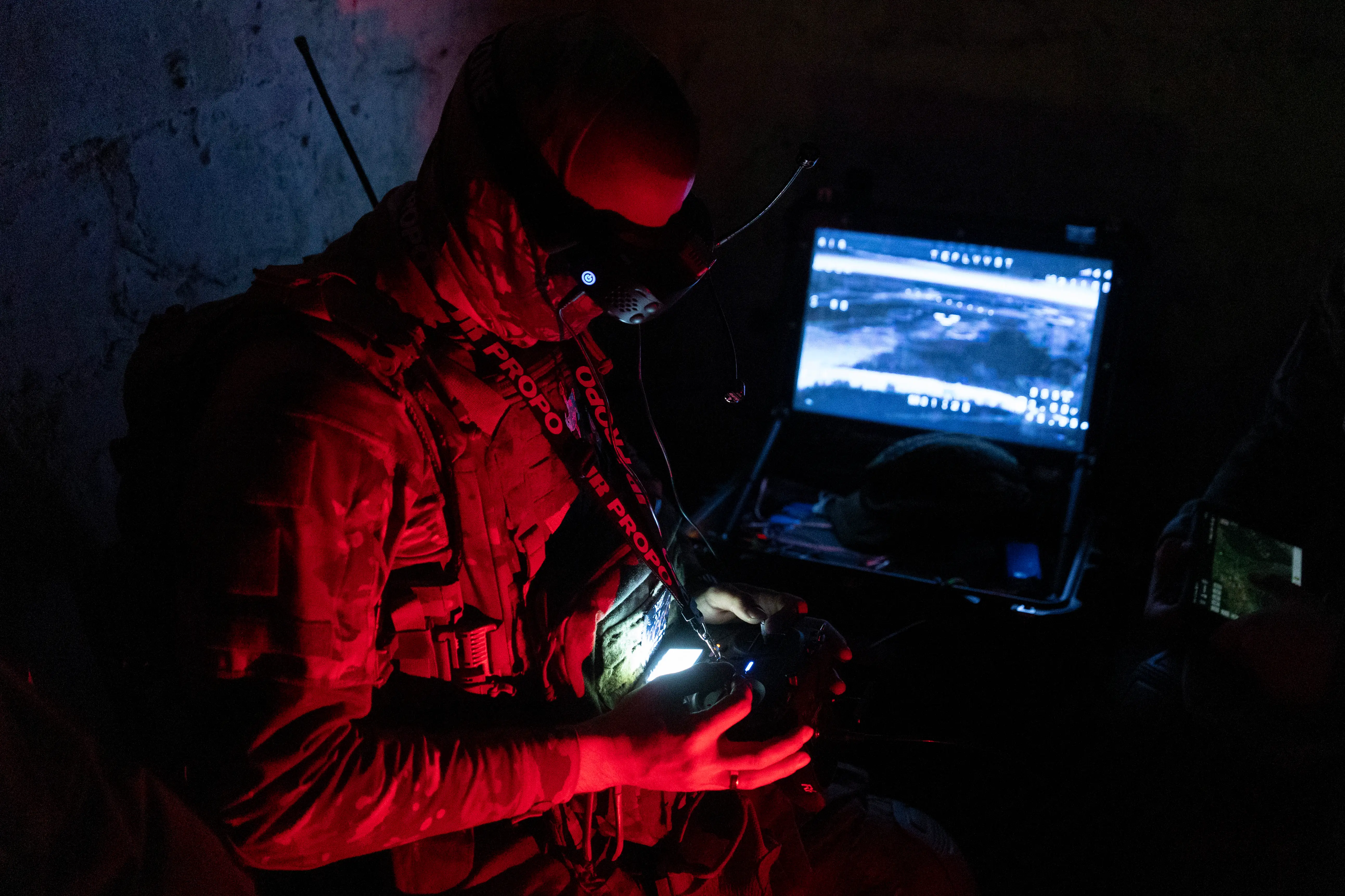 Helmeted person in tactical gear operates a handheld controller beside a laptop in dark red lighting.