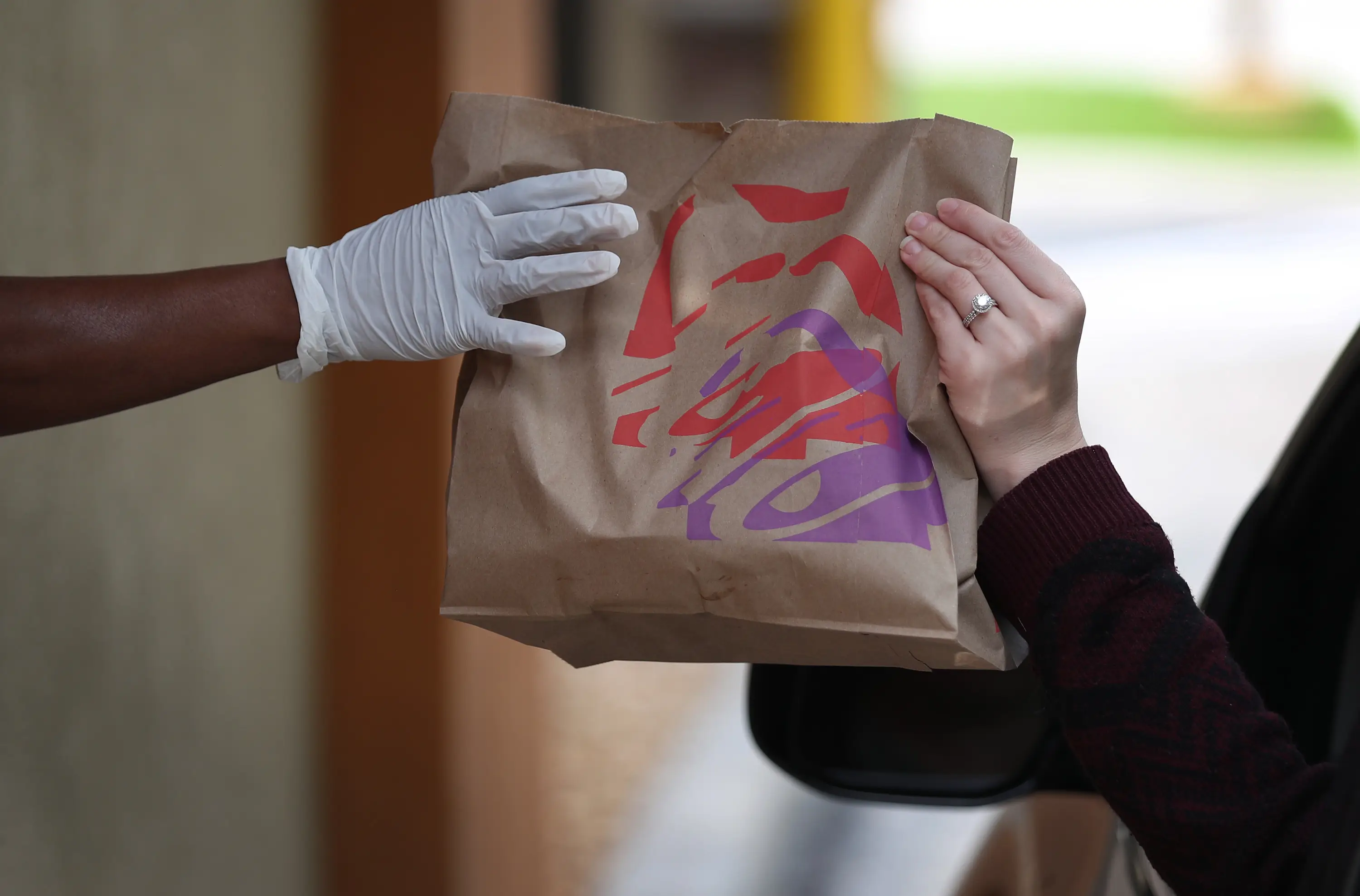 One person wearing a plastic glove hands a bag of Taco Bell food to another person with a ring on their finger.
