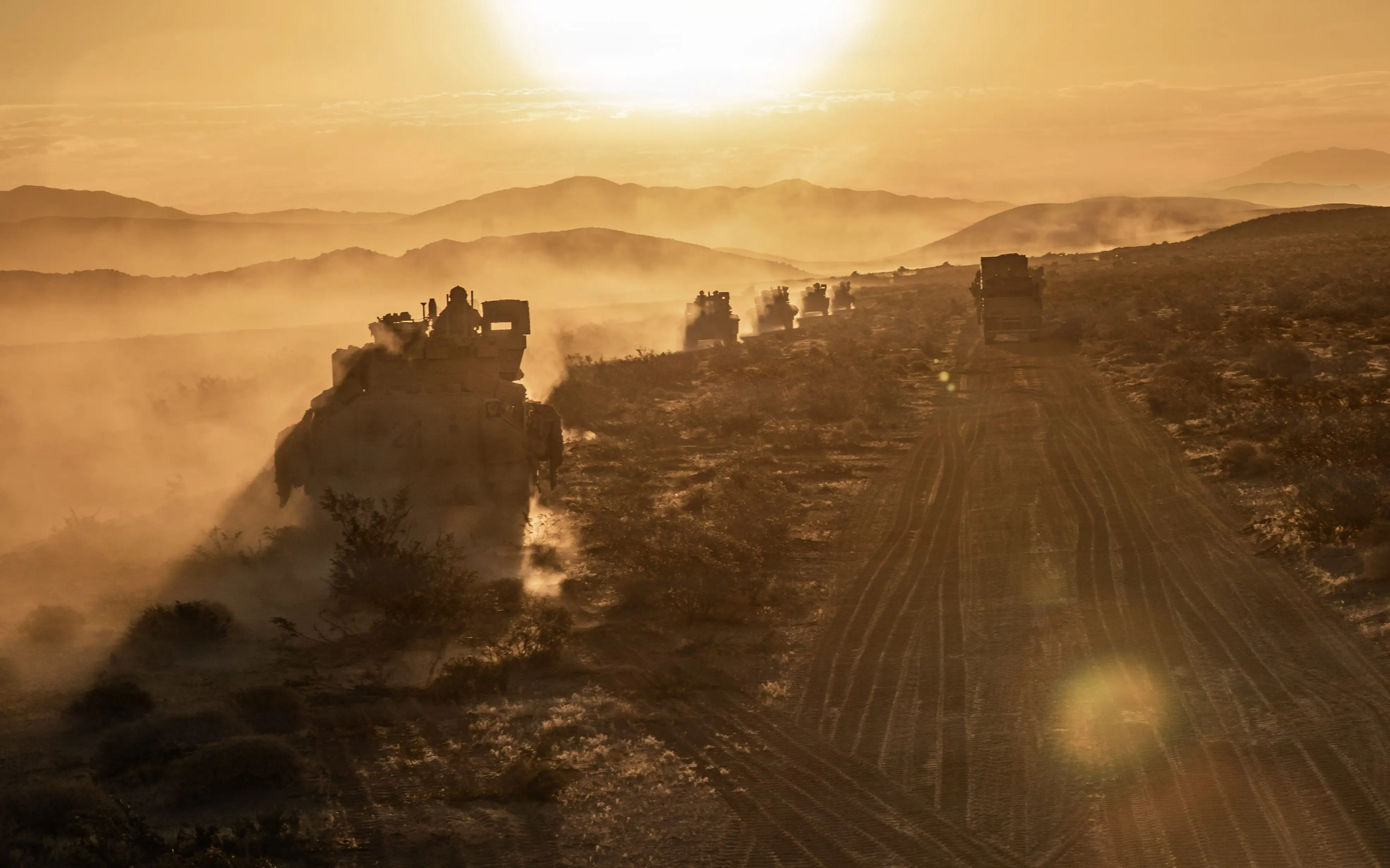 Vehicles move through the desert as the sun is seen on the horizon.
