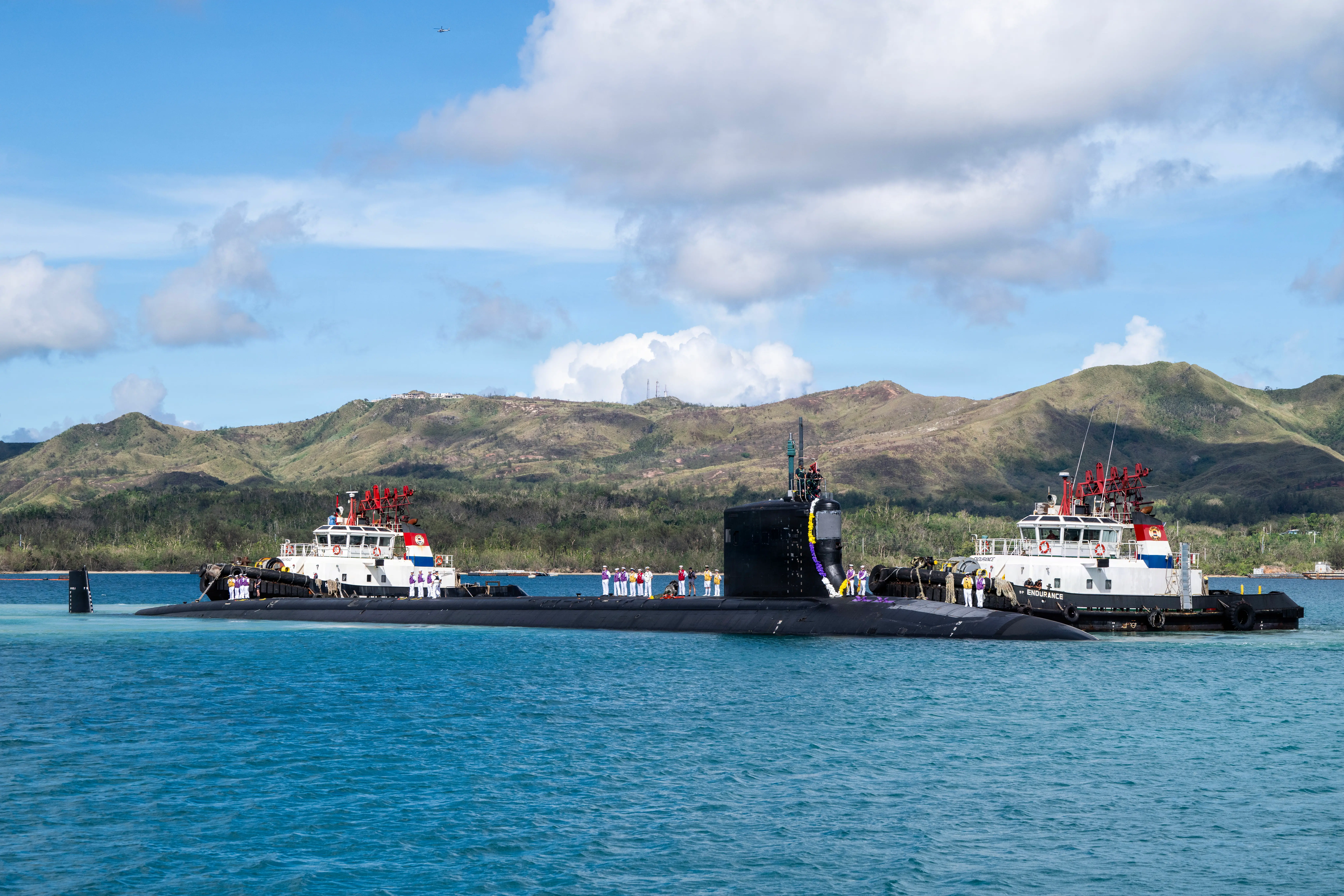 A black submarine sits in blue water surrounded by two boats off a coastline.