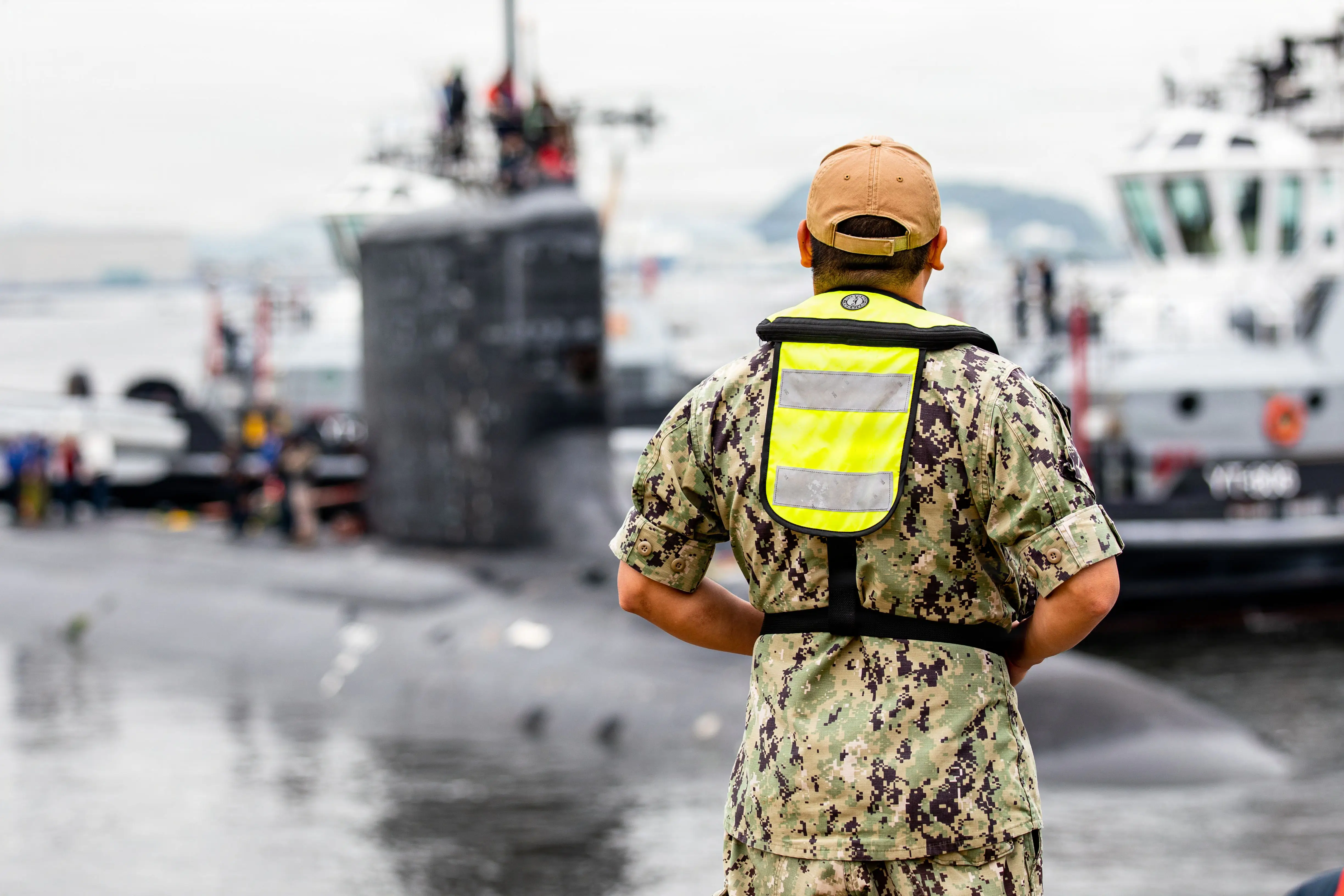 A man wearing camouflage stands in front of a black submarine that's docked near a boat.